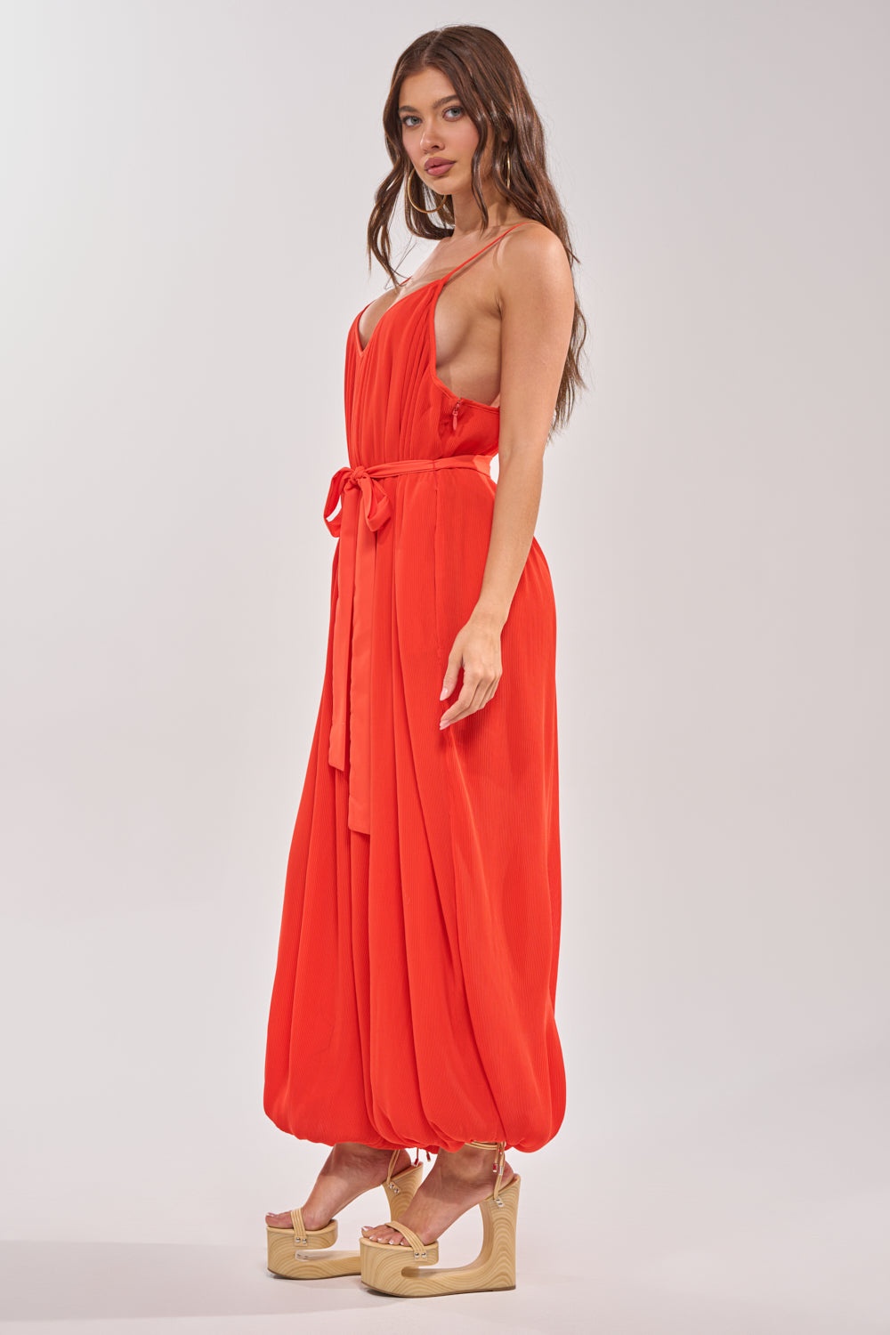 A woman in the VACATIONING PLEATED JUMPSUIT IN ORANGE, featuring a tie belt and balloon hem, stands in beige platform sandals against a plain light background. 2