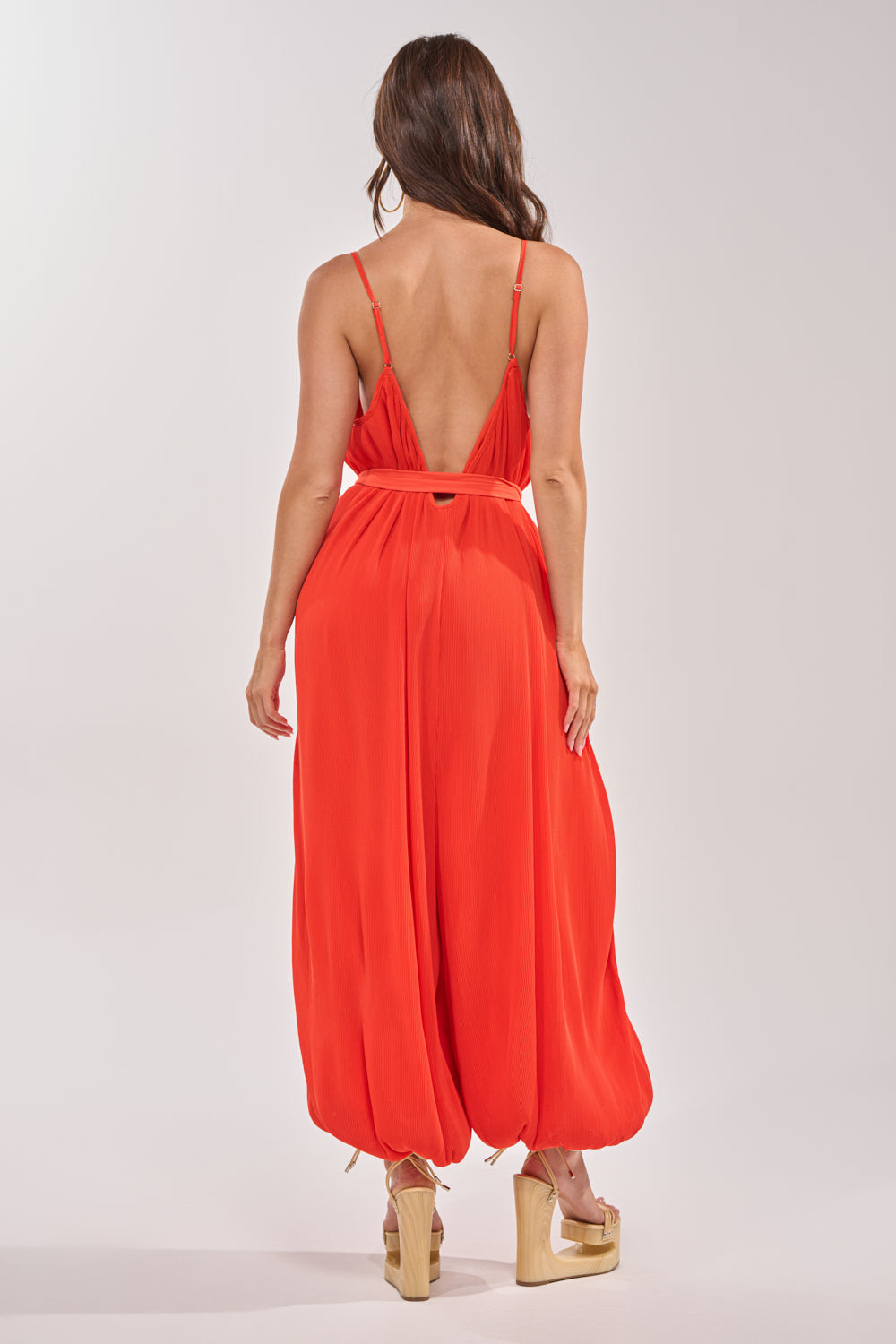 A woman with long brown hair and platform sandals faces away, wearing the VACATIONING PLEATED JUMPSUIT IN ORANGE with a deep V-back and wide, flowing legs. She stands against a plain, light background. 3