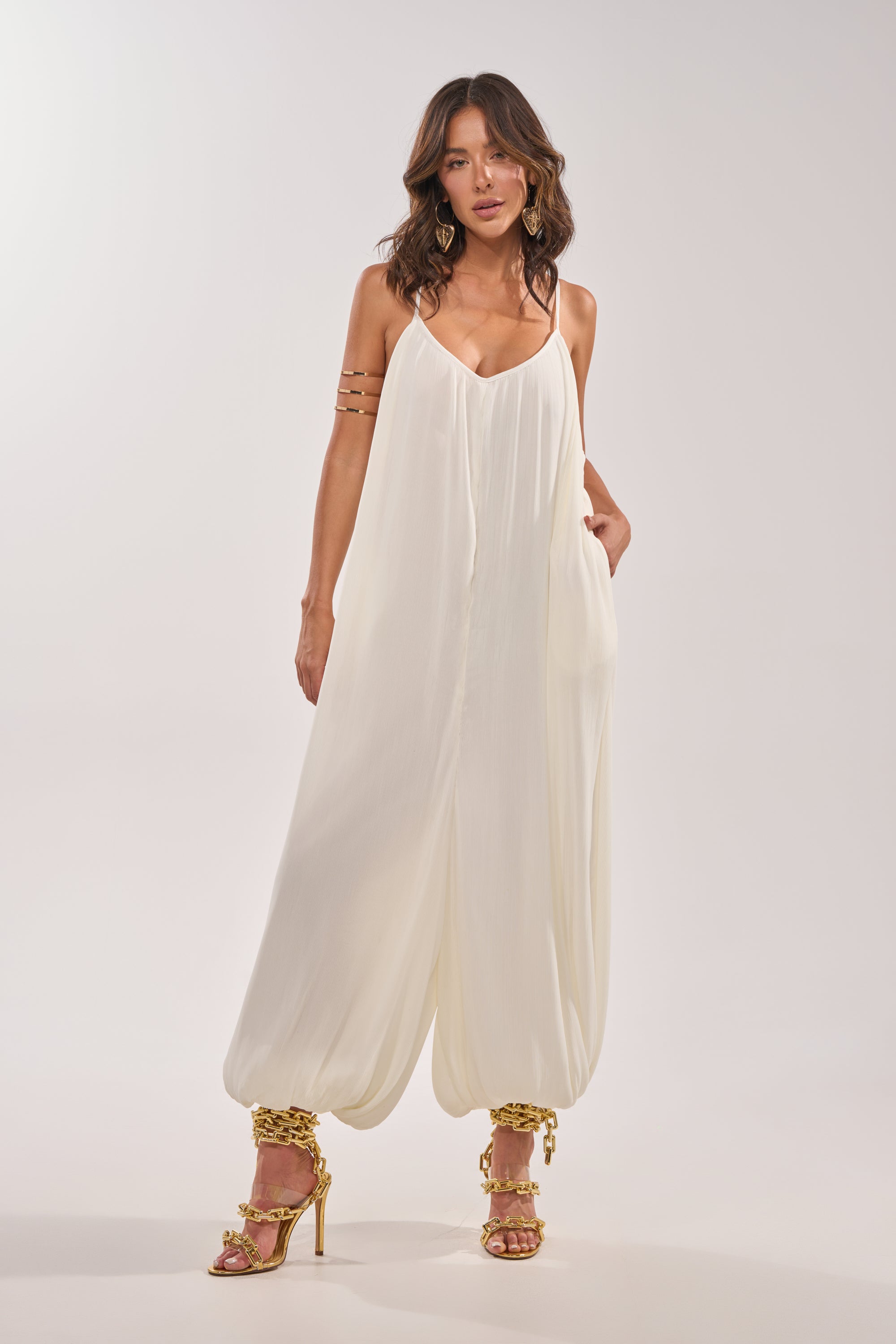 A woman in the HALO SLINKY SLEEVELESS JUMPSUIT with gathered ankles, gold high-heeled sandals, and hoop earrings stands against a plain light background. 0