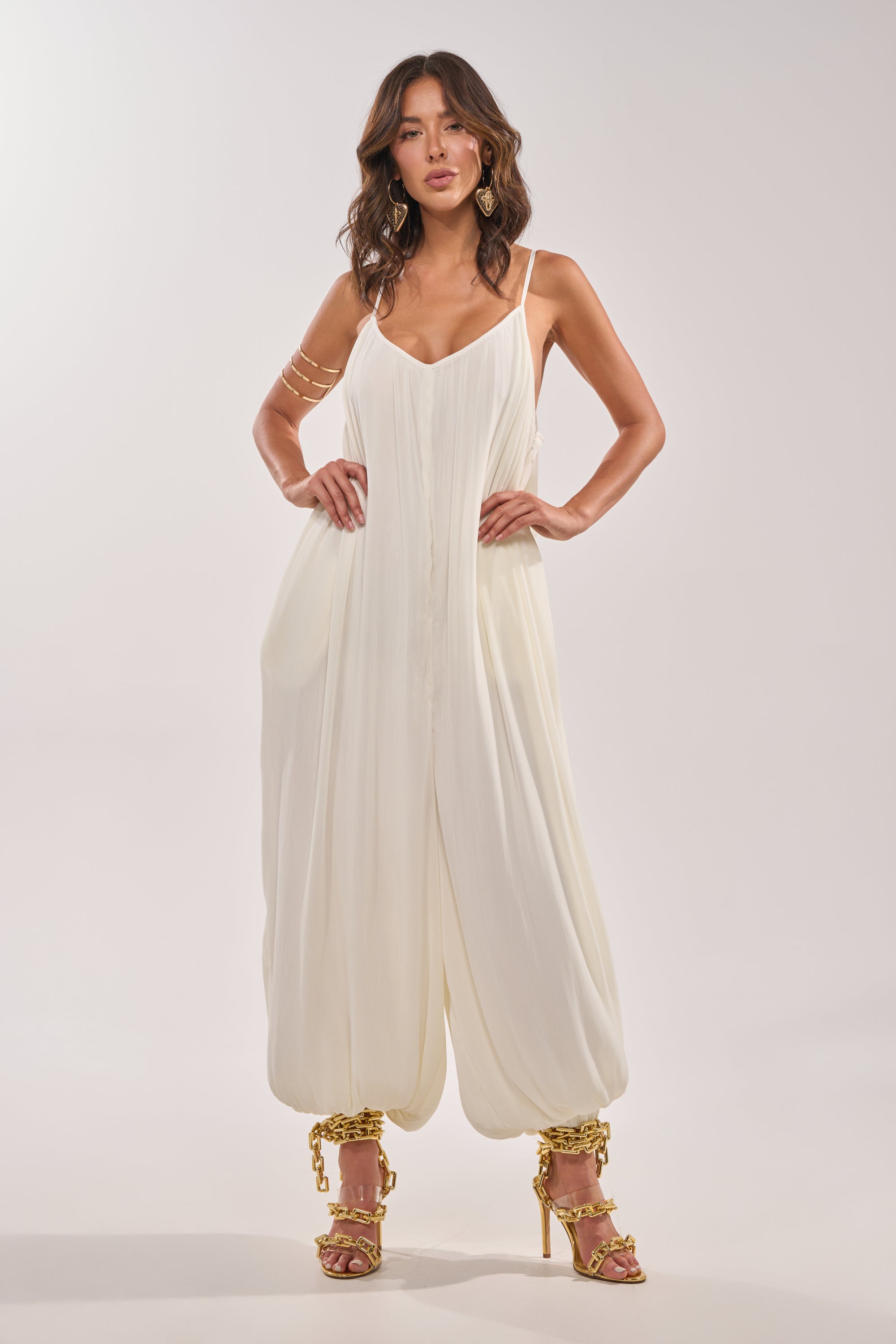 A woman poses in the HALO SLINKY SLEEVELESS JUMPSUIT, featuring loose, gathered ankles. She pairs it with gold strappy heels, gold earrings, and arm cuffs. Her wavy hair is styled loosely for a chic, effortless look against a white background. 2