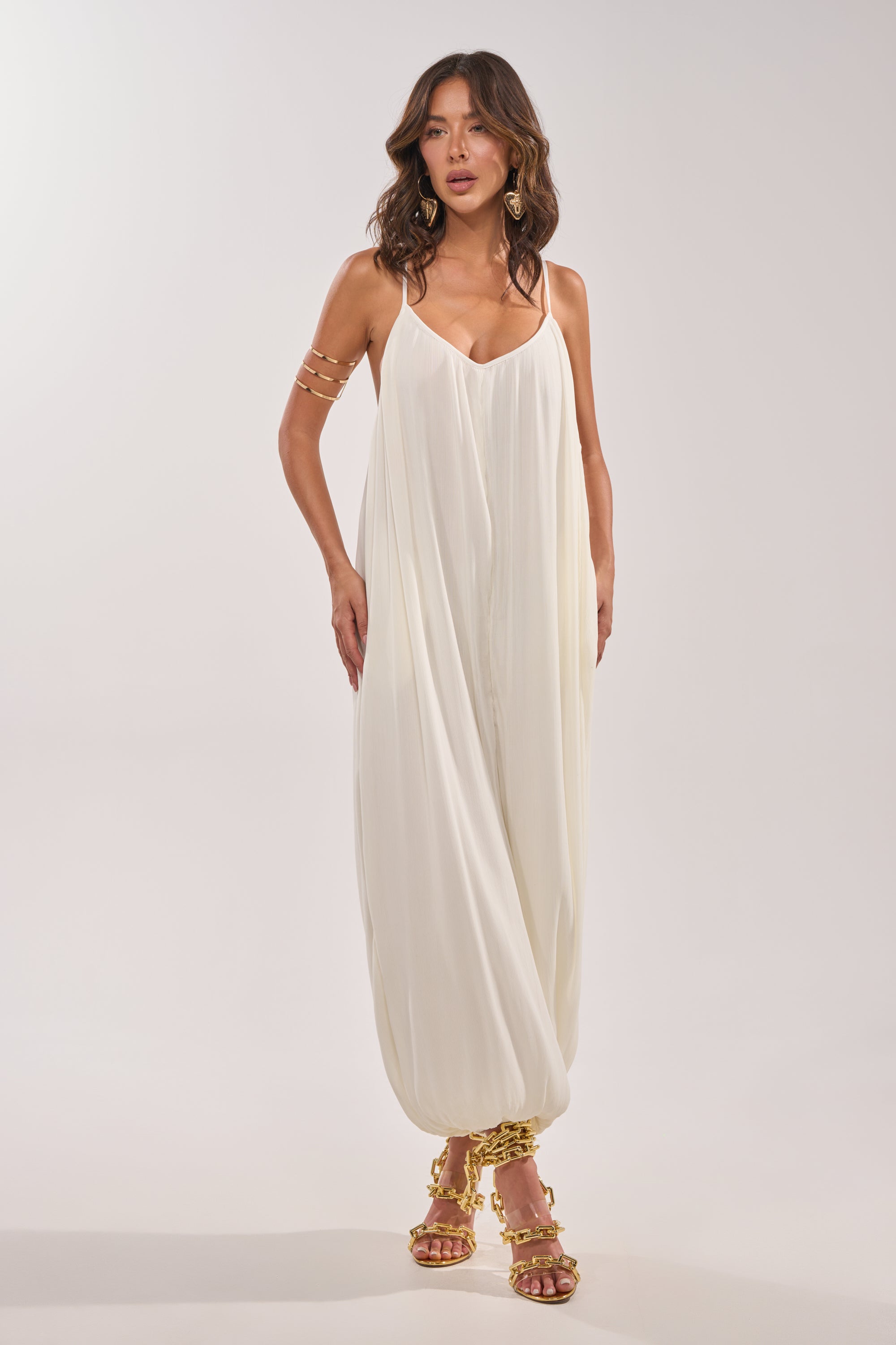 A woman models the HALO SLINKY SLEEVELESS JUMPSUIT, a loose, airy white piece with spaghetti straps, paired with gold statement earrings, a gold arm cuff, and gold strappy sandals against a plain light backdrop. 1
