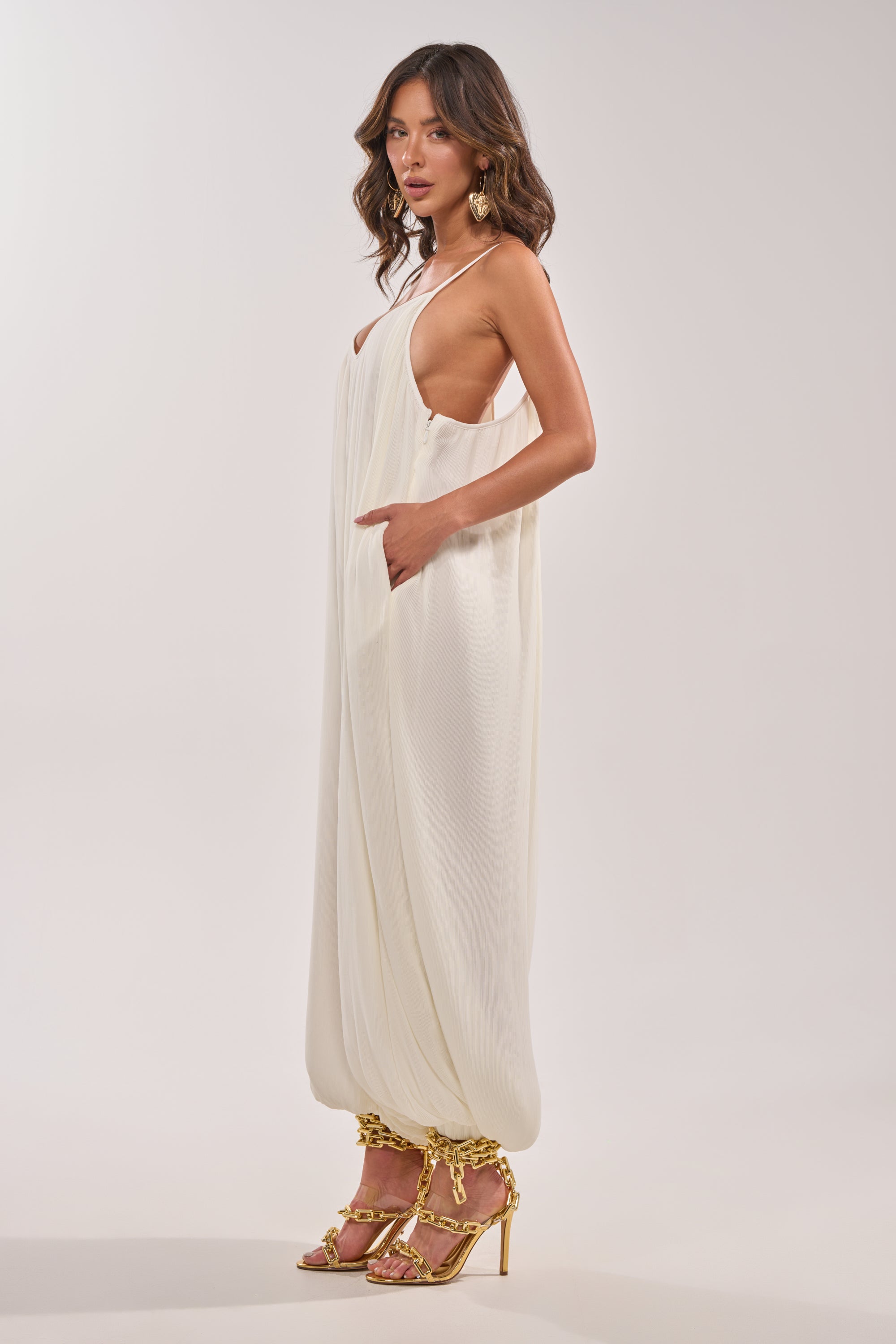 A woman poses sideways in the HALO SLINKY SLEEVELESS JUMPSUIT, featuring low-cut sides and gathered ankles, styled with gold chain-embellished heels and large hoop earrings against a plain light background. 3