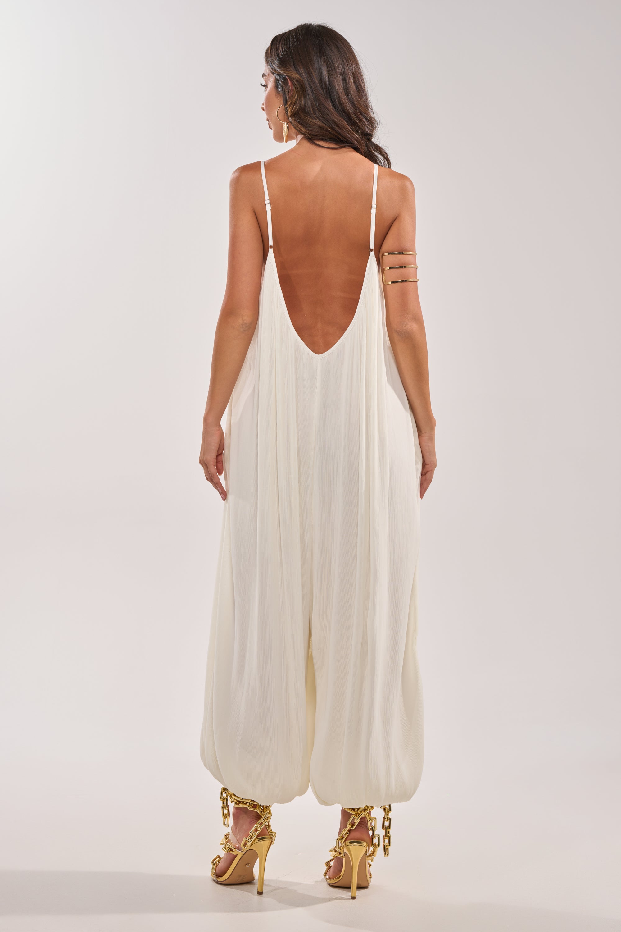A woman stands with her back to the camera, wearing the HALO SLINKY SLEEVELESS JUMPSUIT in white with a low scoop back and gathered ankles, paired with gold high-heeled sandals. 4