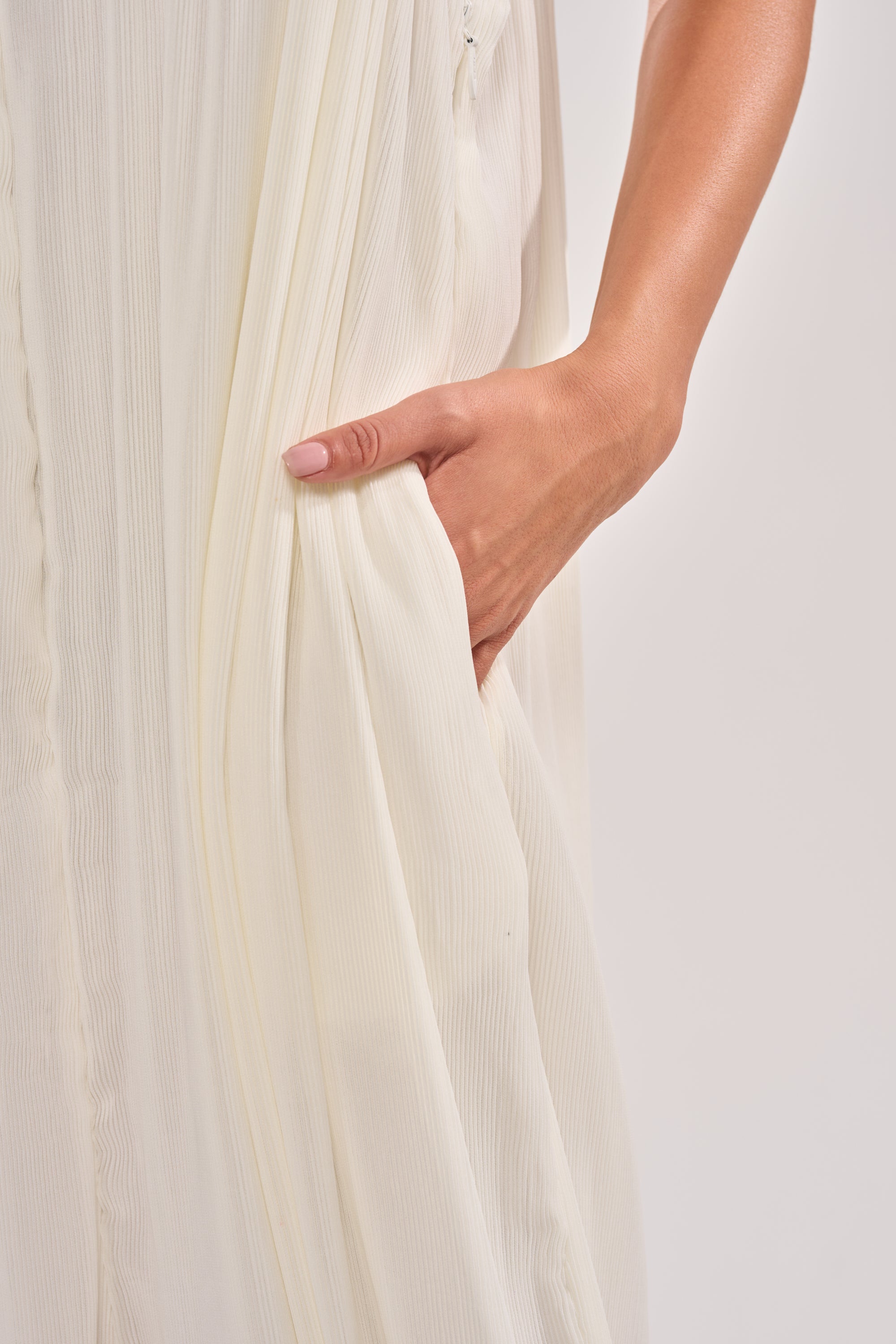 A hand with light pink nail polish rests in the pocket of the flowing, pleated, off-white HALO SLINKY SLEEVELESS JUMPSUIT. 5
