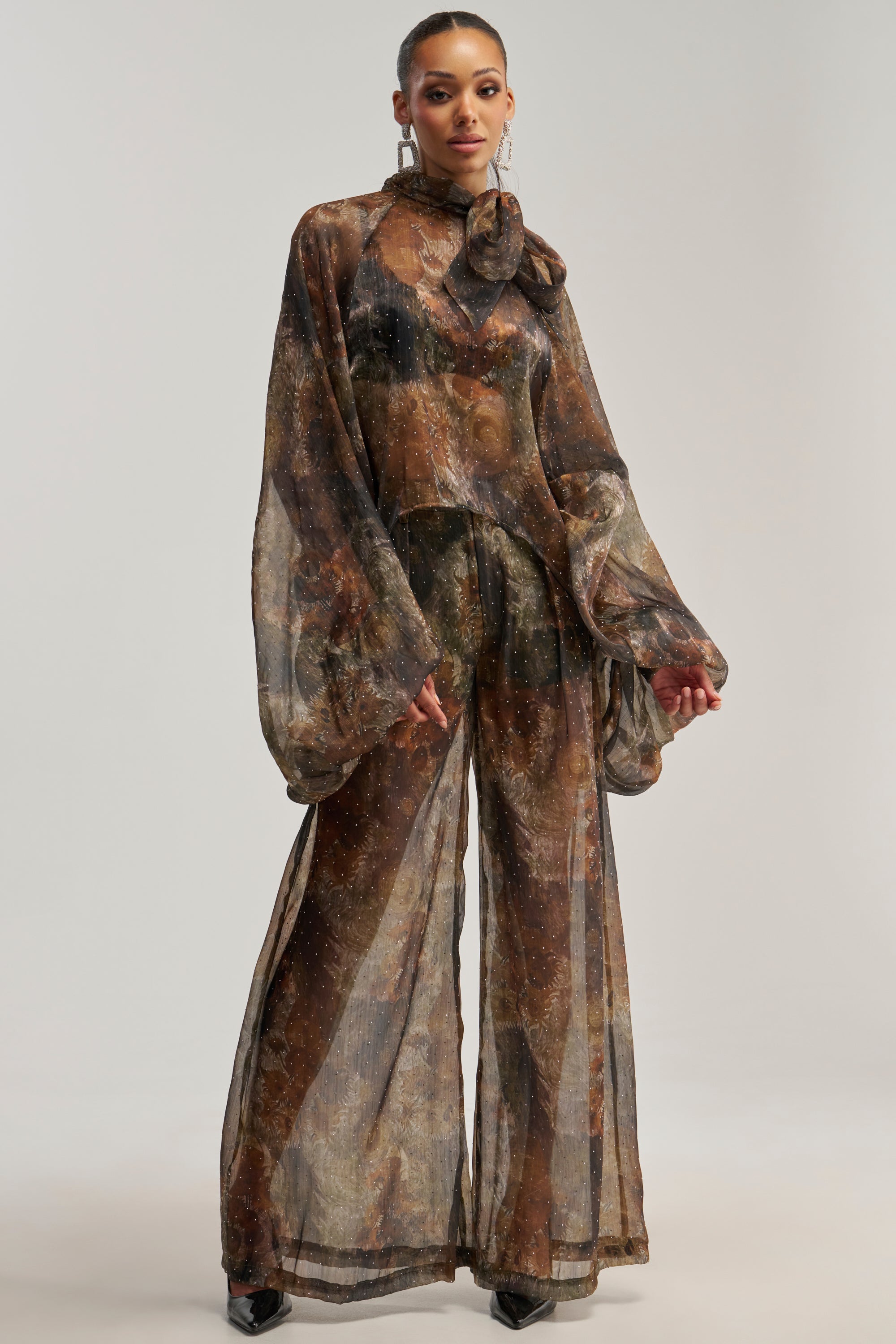 A person wearing the LADY LUXE TROUSER—a pair of flowing green floral chiffon pants—styled with a sheer, long-sleeve, high-neck top featuring an earthy abstract pattern and a bow. Their hair is tied back and accessories are kept minimal to complement the look.