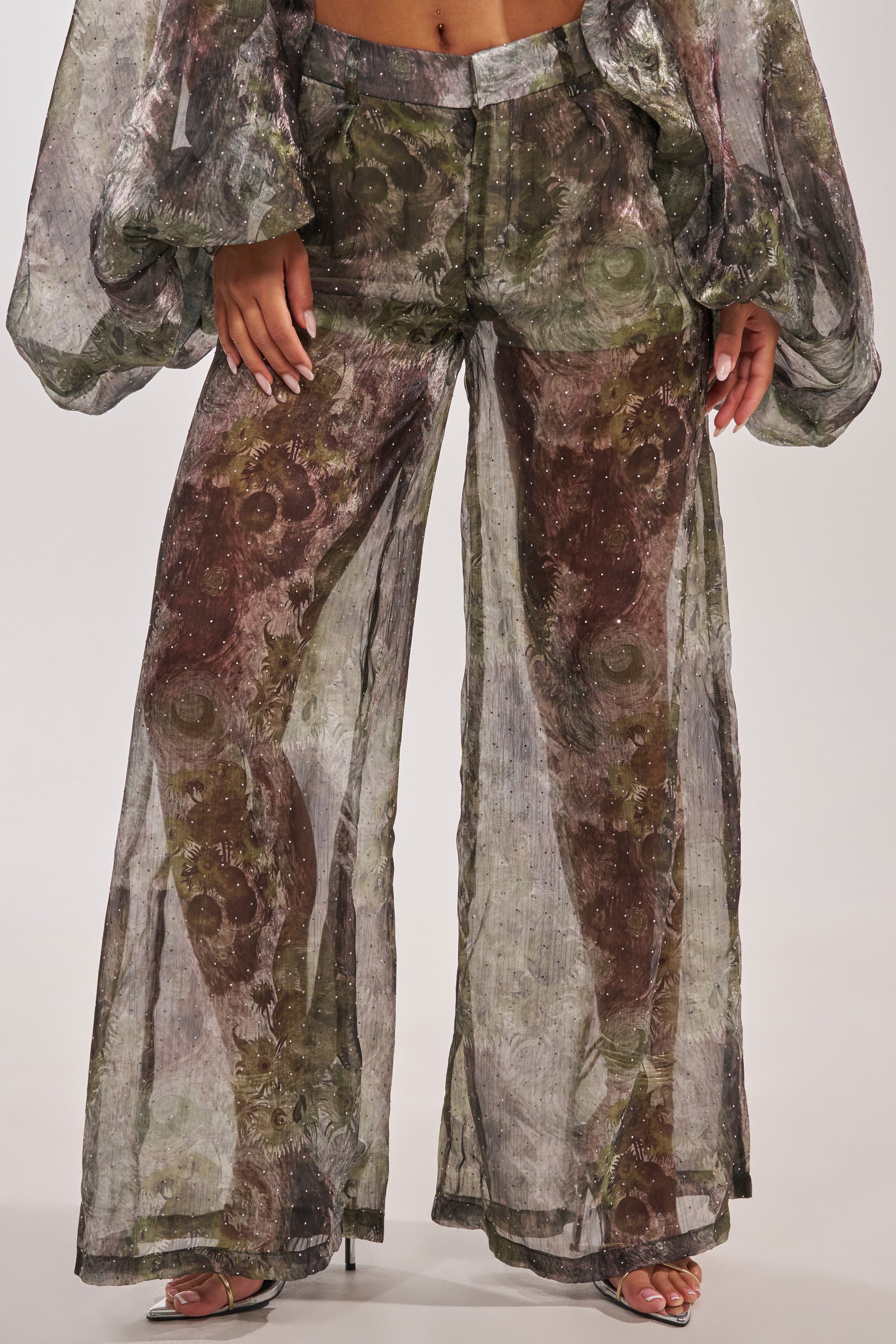 A person wearing the MYSTIQUE FLORAL CHIFFON PANT—high-waisted, sheer, wide-leg pants with a green and brown abstract pattern—paired with a matching long-sleeve top and heeled sandals on a light background. 2