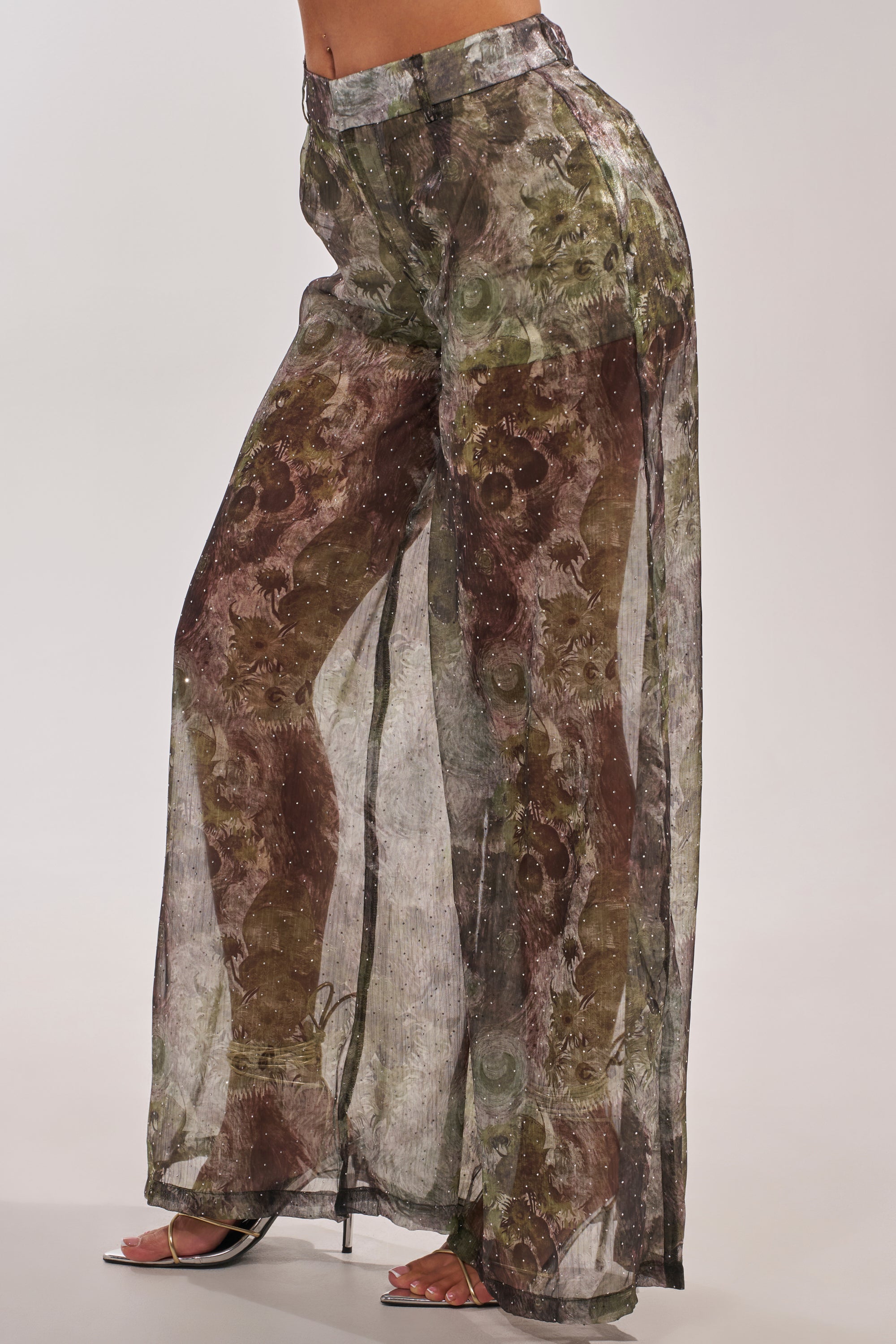 A person models the MYSTIQUE FLORAL CHIFFON PANT featuring a high-waisted abstract green and brown floral design, styled with open-toe heels, set against a simple light background. 3