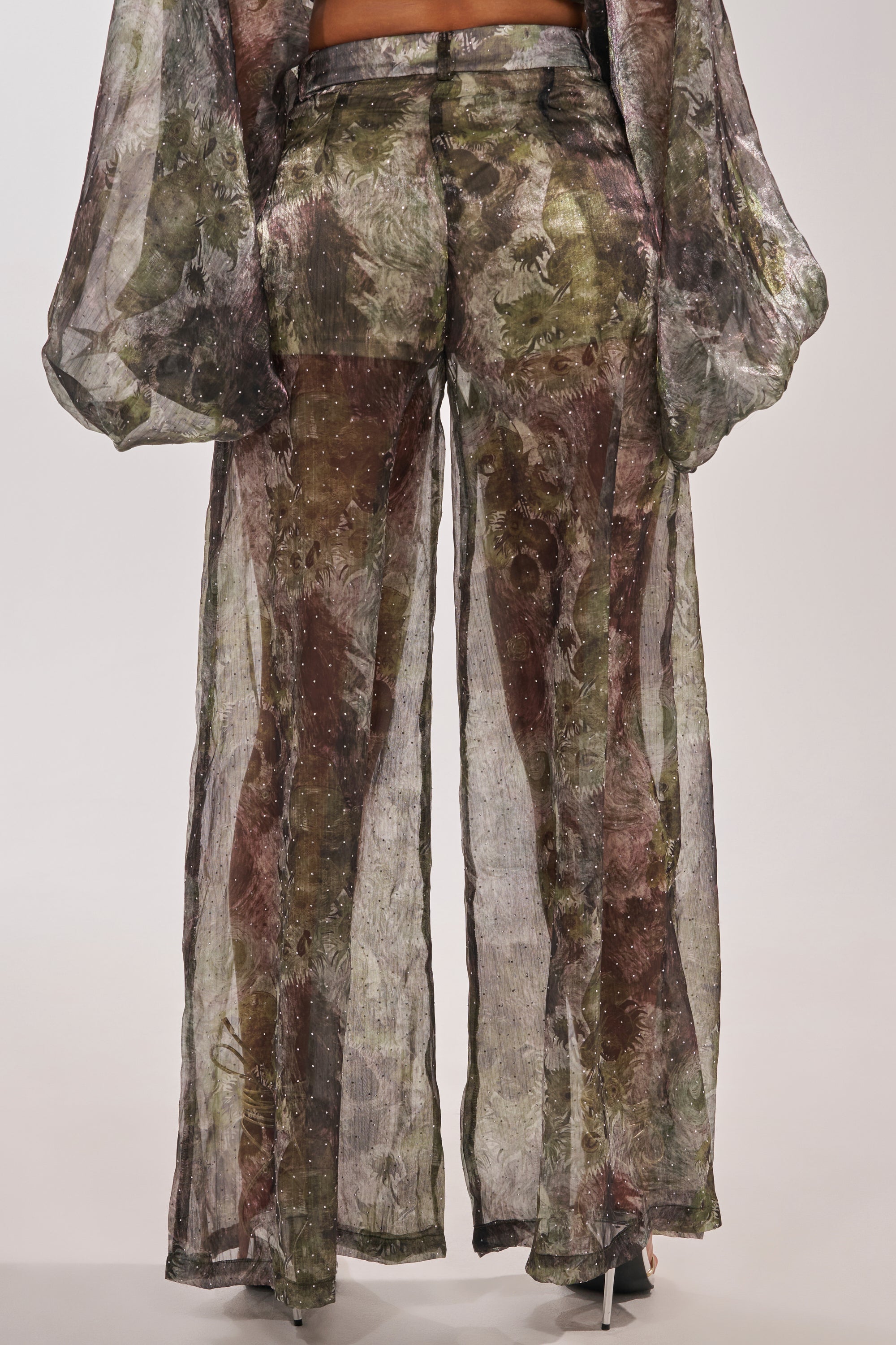 Wearing the MYSTIQUE FLORAL CHIFFON PANT—high-waisted, wide-leg pants with a subtle shimmer, sheer chiffon overlay, and matching semi-transparent long sleeves. The partially see-through design reveals fitted shorts underneath. 4