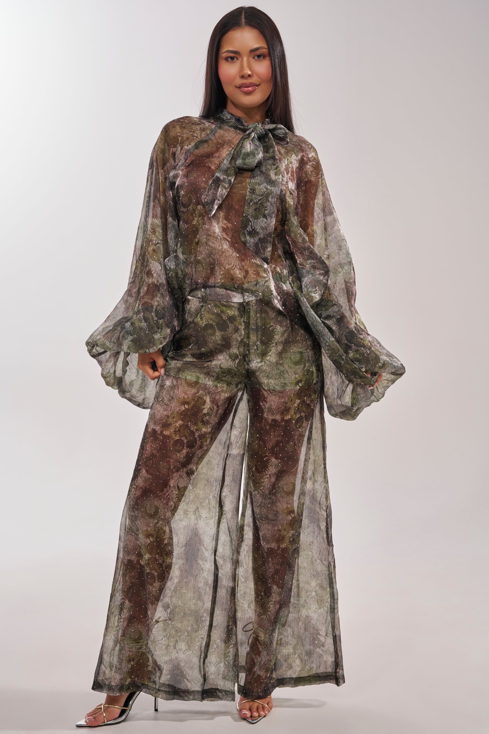 A woman models the MYSTIQUE FLORAL CHIFFON PANT with sheer wide legs, long bell sleeves, a statement neck tie, and heels against a plain light backdrop. 5