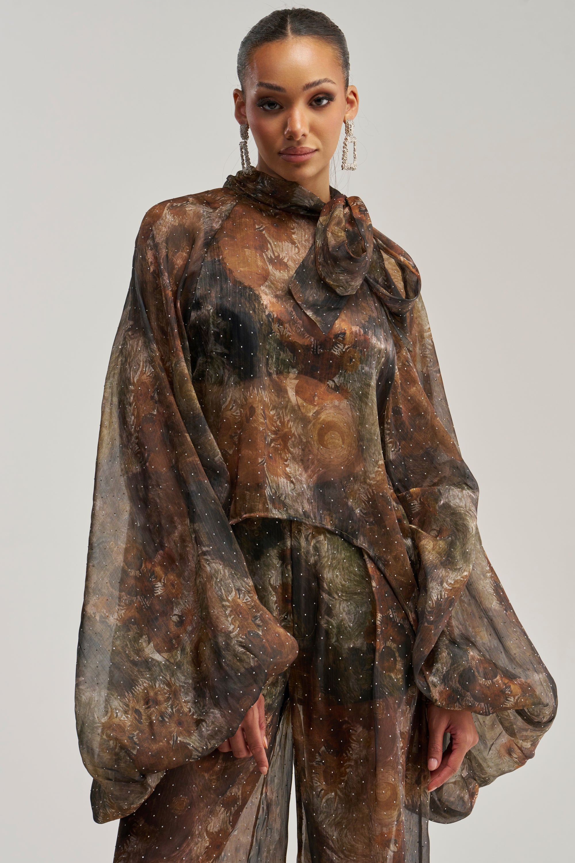 A woman wearing the LADY LUXE BLOUSE—a sheer, long-sleeved brown and black patterned piece featuring a high neck and oversized bow, reminiscent of elegant balloon sleeves—paired with matching wide-leg pants and statement earrings, poses against a plain light background.
