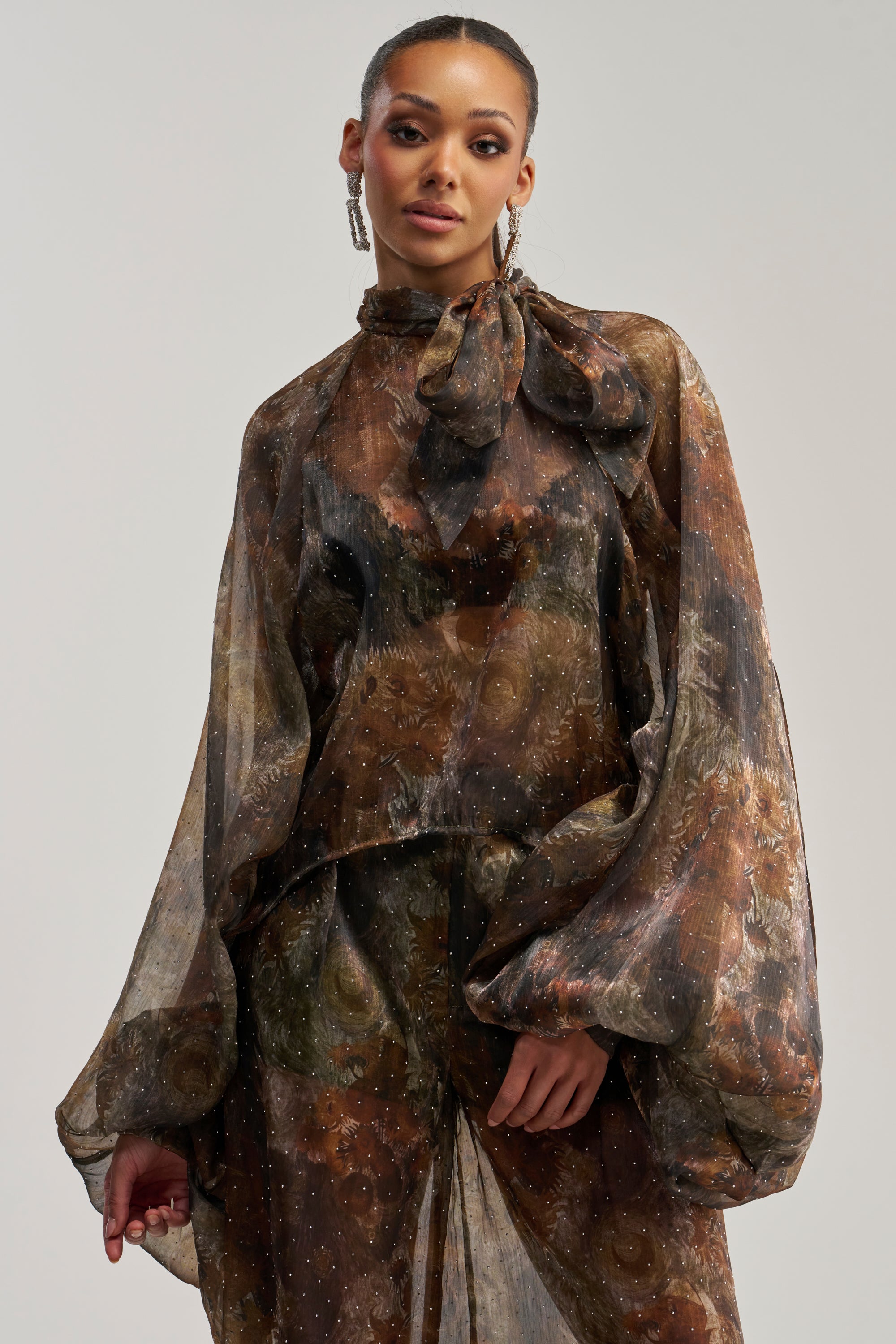 Wearing the LADY LUXE BLOUSE, a sheer brown floral design featuring a neck bow and voluminous balloon sleeves, the woman pairs it with a matching skirt. She stands against a light, neutral background, gazing confidently at the camera.