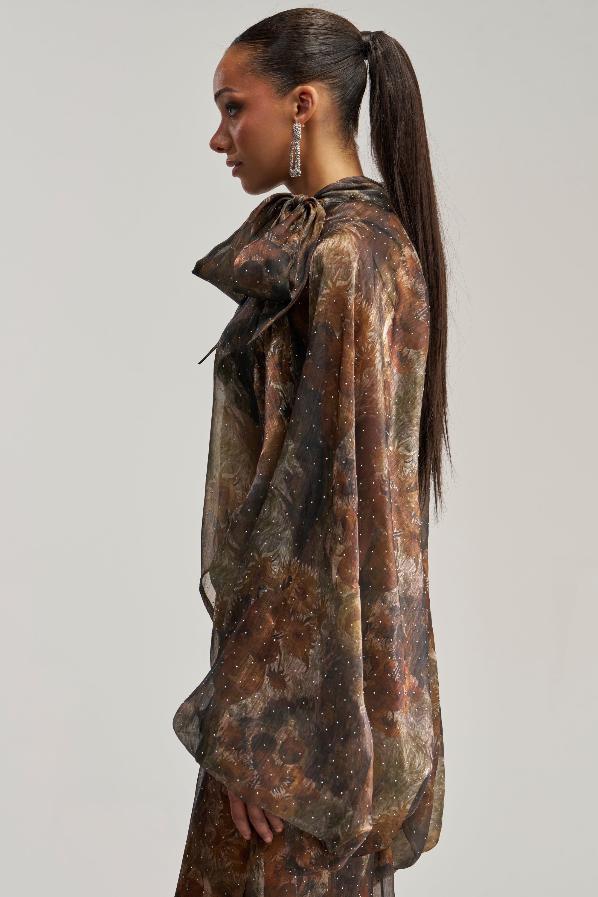 A woman with a sleek ponytail stands in profile against a plain light background, wearing the LADY LUXE BLOUSE—a sheer brown floral top featuring long sleeves and a statement large bow at the neck.