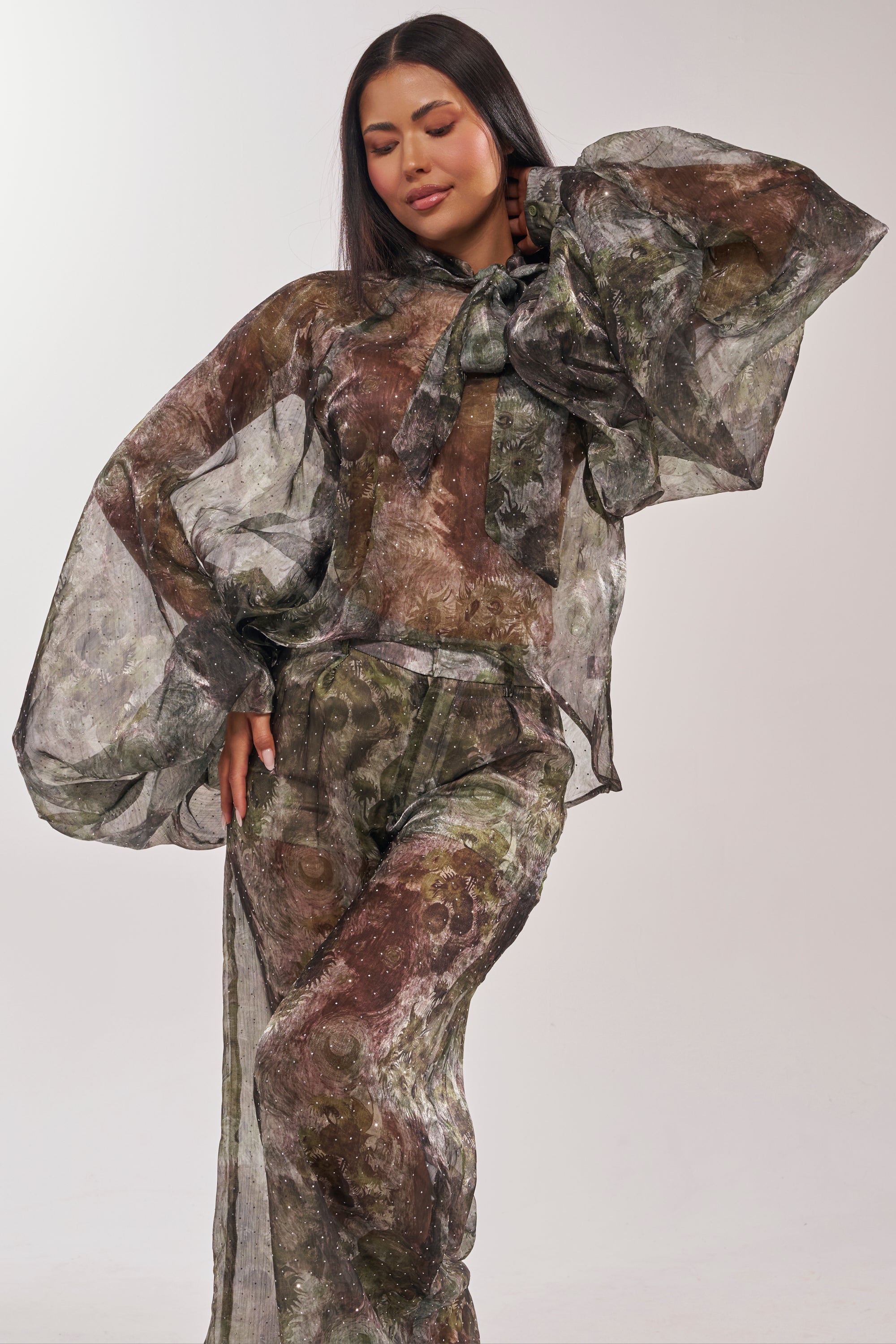 A woman models the MYSTIQUE FLORAL CHIFFON BLOUSE, featuring sheer balloon sleeves, earth-toned floral patterns, and a large neck bow. She poses with one hand on her hip and smiles gently against a light background. 0
