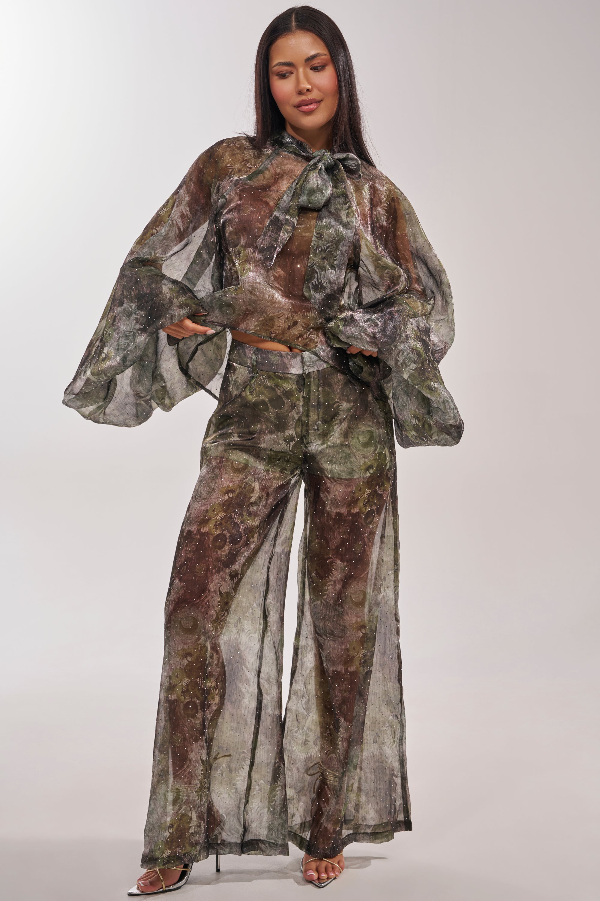 A woman stands smiling in the MYSTIQUE FLORAL CHIFFON BLOUSE, featuring sheer balloon sleeves, a high-neck bow, subtle rhinestone accents, and wide-leg pants, against a plain background. 9