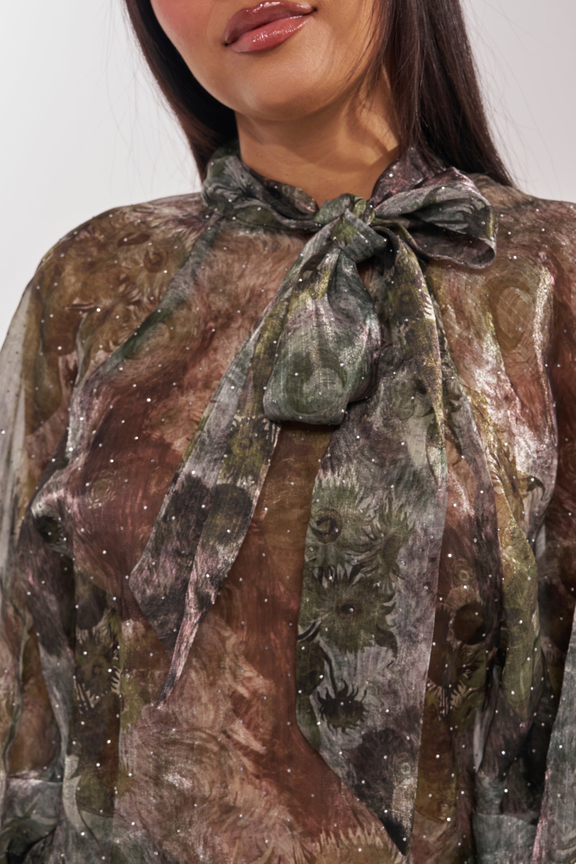 A close-up of a woman wearing the MYSTIQUE FLORAL CHIFFON BLOUSE, featuring sheer fabric, balloon sleeves, a nature-inspired abstract print in earthy tones, sparkling accents, and a statement bow at the high neckline. 10