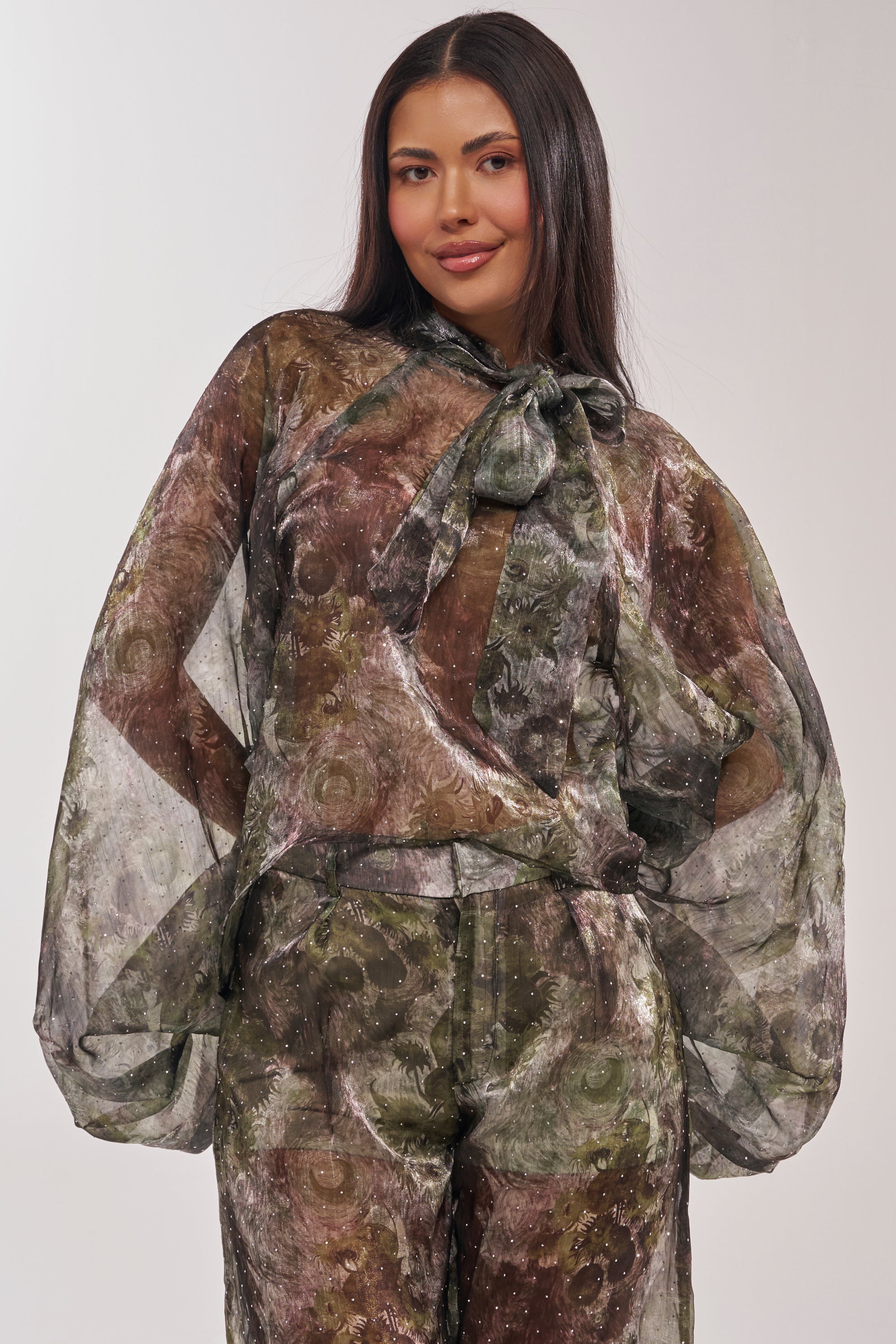 A woman with long dark hair smiles softly while wearing the MYSTIQUE FLORAL CHIFFON BLOUSE, featuring sheer floral fabric, a neck tie, and voluminous sleeves, standing against a plain light background. 2