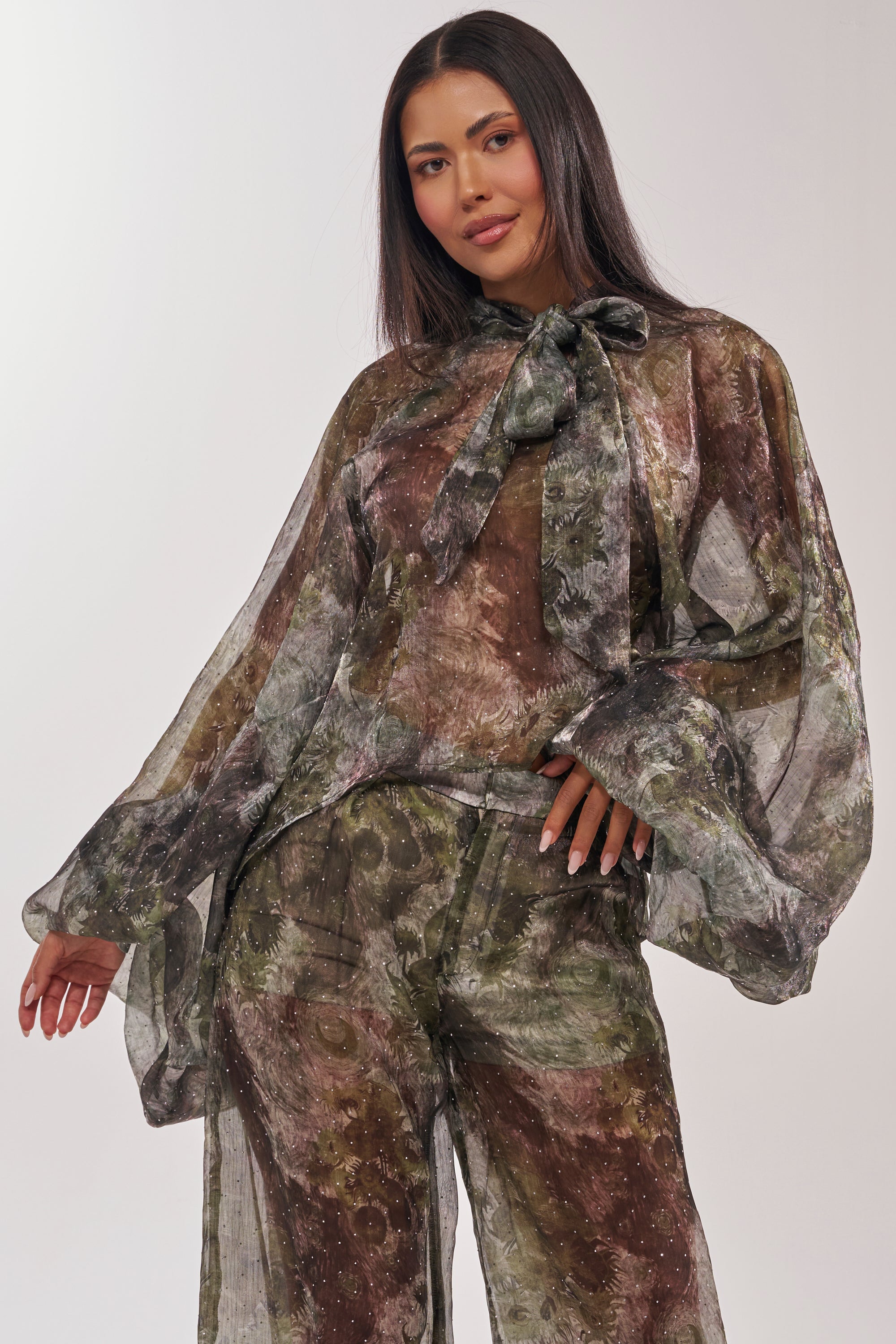 A woman with long, straight dark hair models the MYSTIQUE FLORAL CHIFFON BLOUSE—featuring a sheer, floral design with brown and green abstract patterns, wide sleeves, and a statement neck bow—against a plain light background. 3