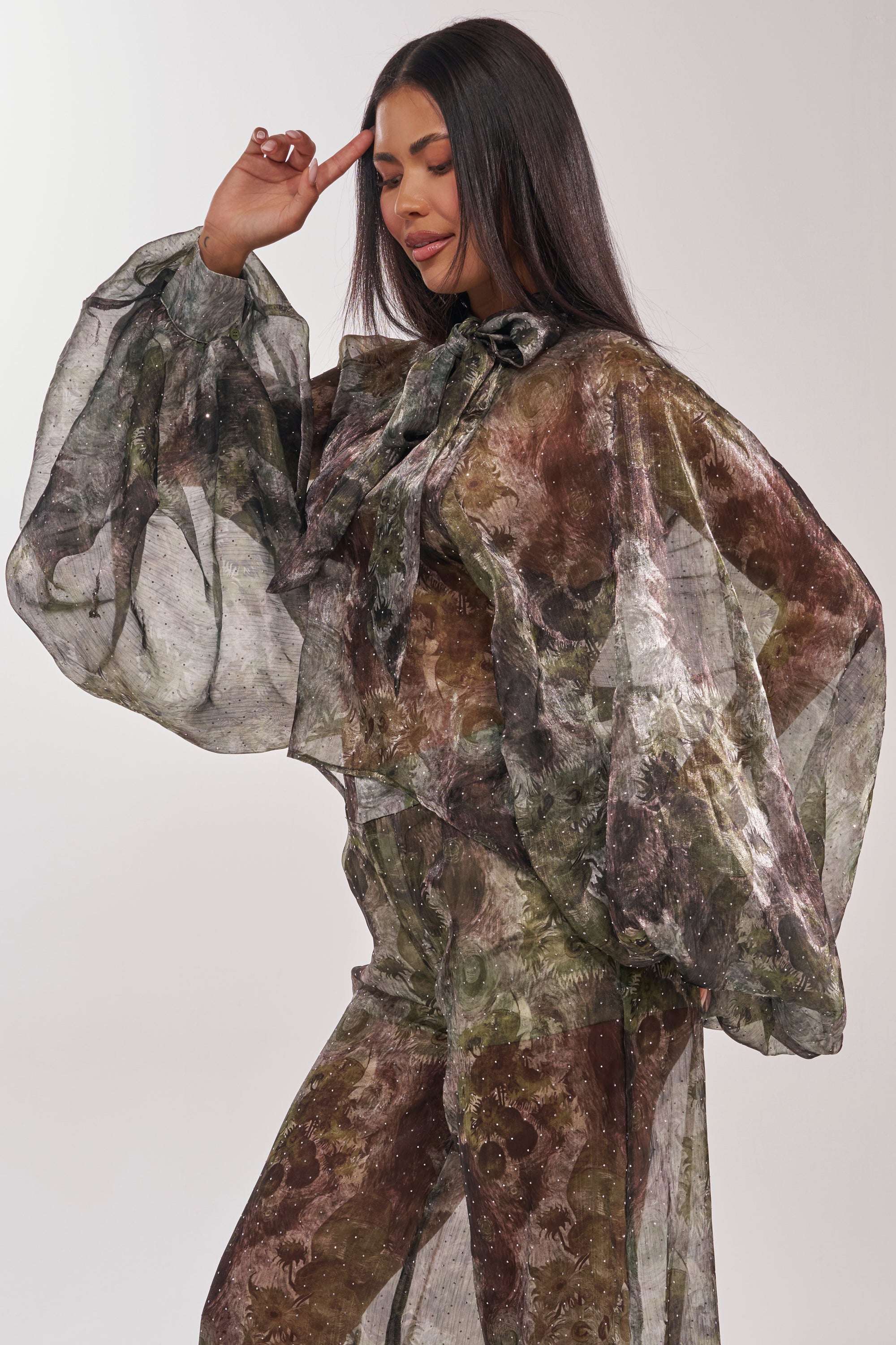 A woman poses with one hand raised to her forehead, wearing the MYSTIQUE FLORAL CHIFFON BLOUSE featuring sheer fabric, voluminous balloon sleeves, a tied neck detail, and earthy floral tones against a plain background. 4