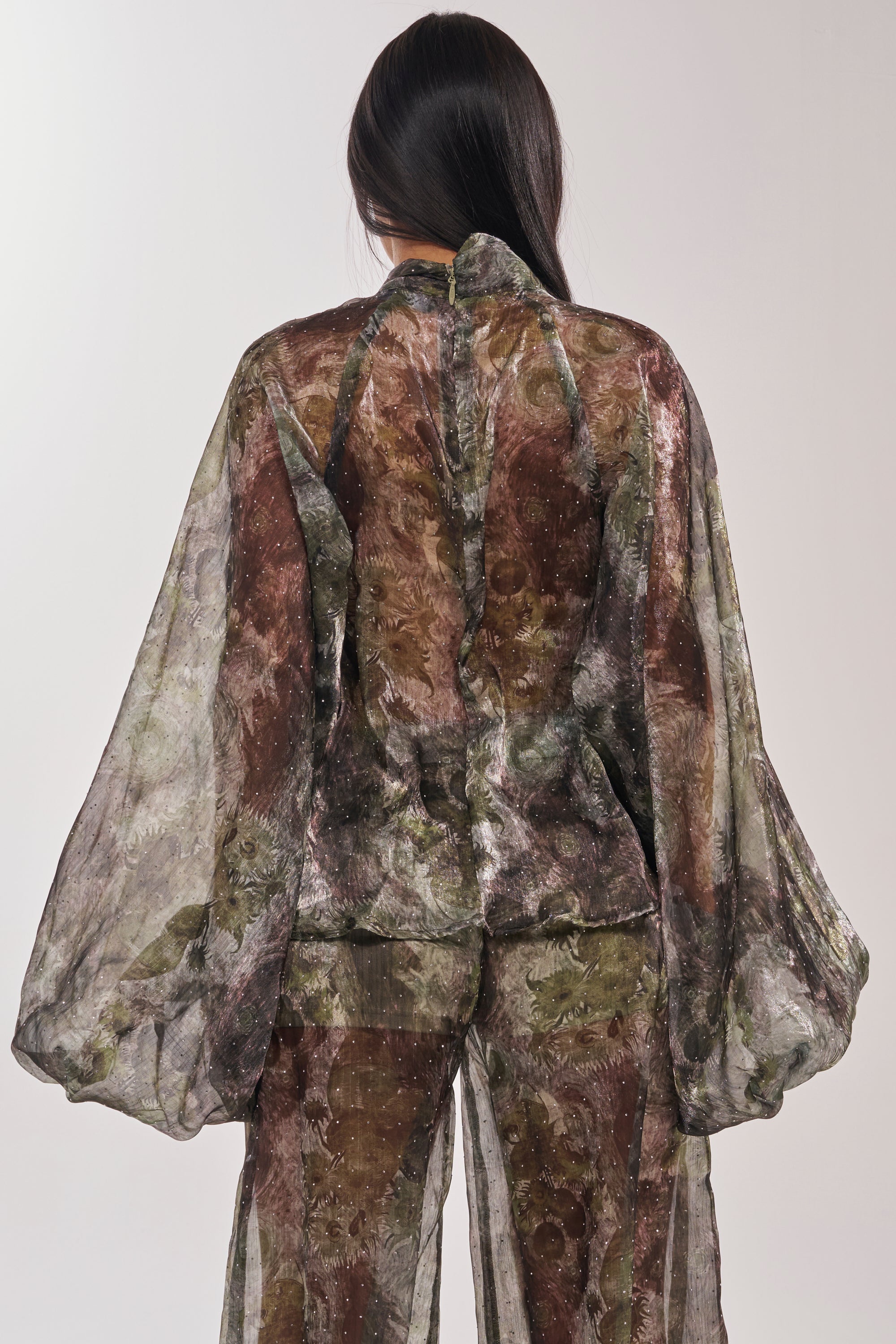 A person with long dark hair faces away, wearing the MYSTIQUE FLORAL CHIFFON BLOUSE in earthy tones with voluminous sleeves and a subtle floral pattern. The background is plain and light-colored. 5