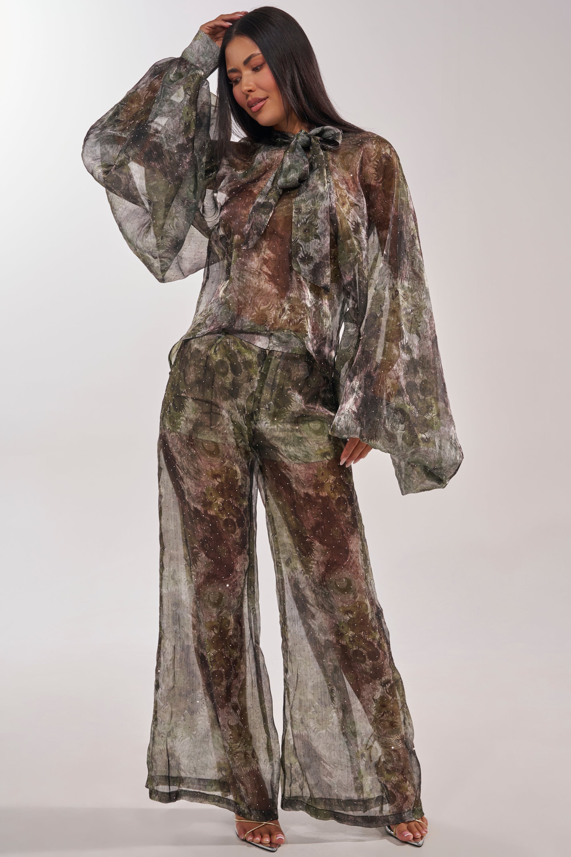 A woman poses with one hand on her head, wearing the MYSTIQUE FLORAL CHIFFON BLOUSE—a sheer floral top with a bow at the collar—paired with matching green and brown abstract wide-leg pants. 6