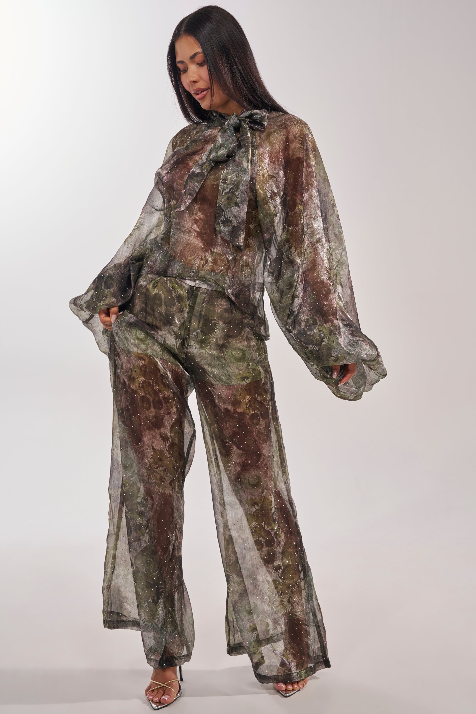 A woman wears the MYSTIQUE FLORAL CHIFFON BLOUSE—a sheer, high-neck, balloon-sleeve top with a large neck bow—paired with matching wide-leg pants in an abstract green and brown print. She stands holding her pants and looking down. 8
