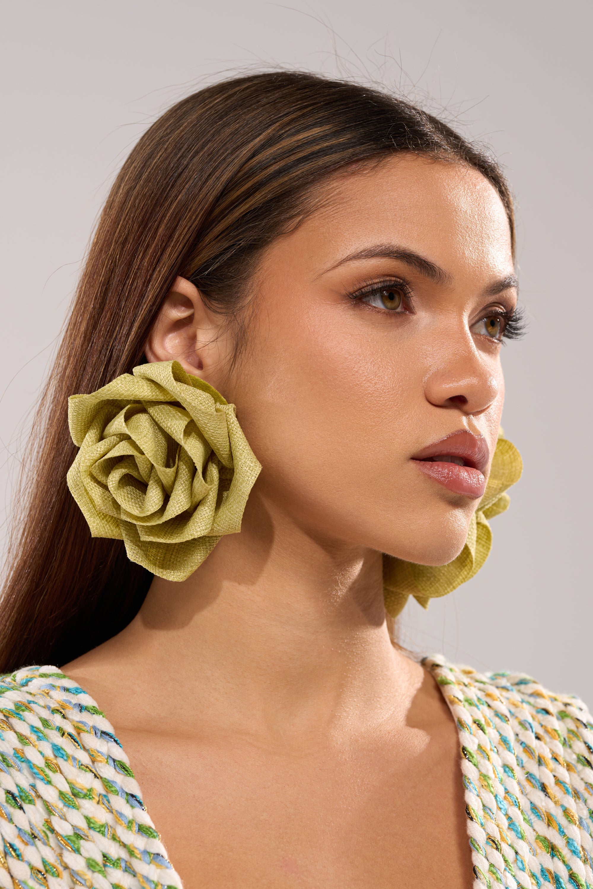 A woman with straight brown hair wears the AT BRUNCH EARRING—large, woven textured floral earrings in light green—and a multicolored top. She gazes to the side with a neutral expression against a plain background. 1