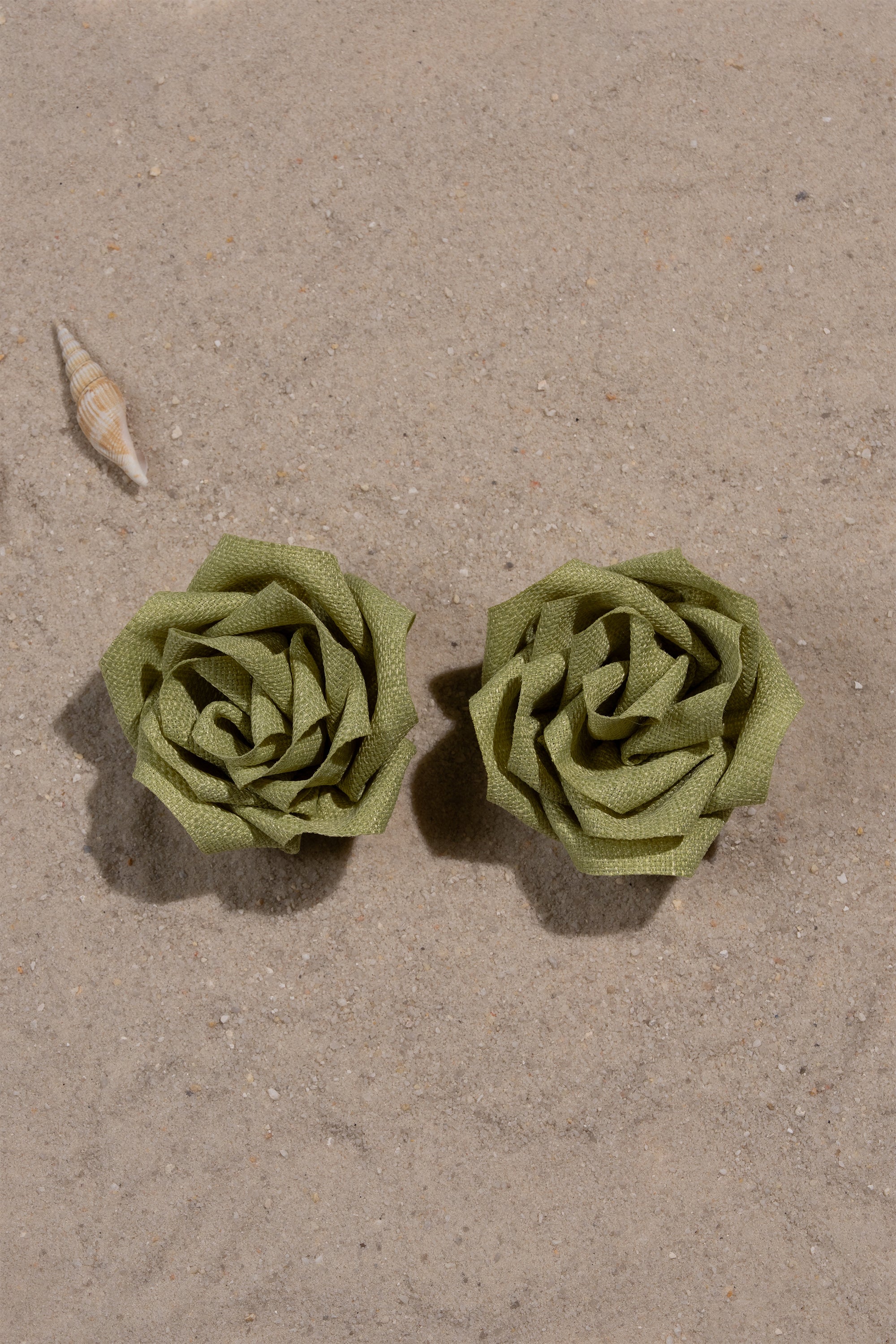 Two AT BRUNCH EARRING statement studs, green and flower-shaped from folded material, are placed side by side on sandy ground with a small seashell nearby. 0