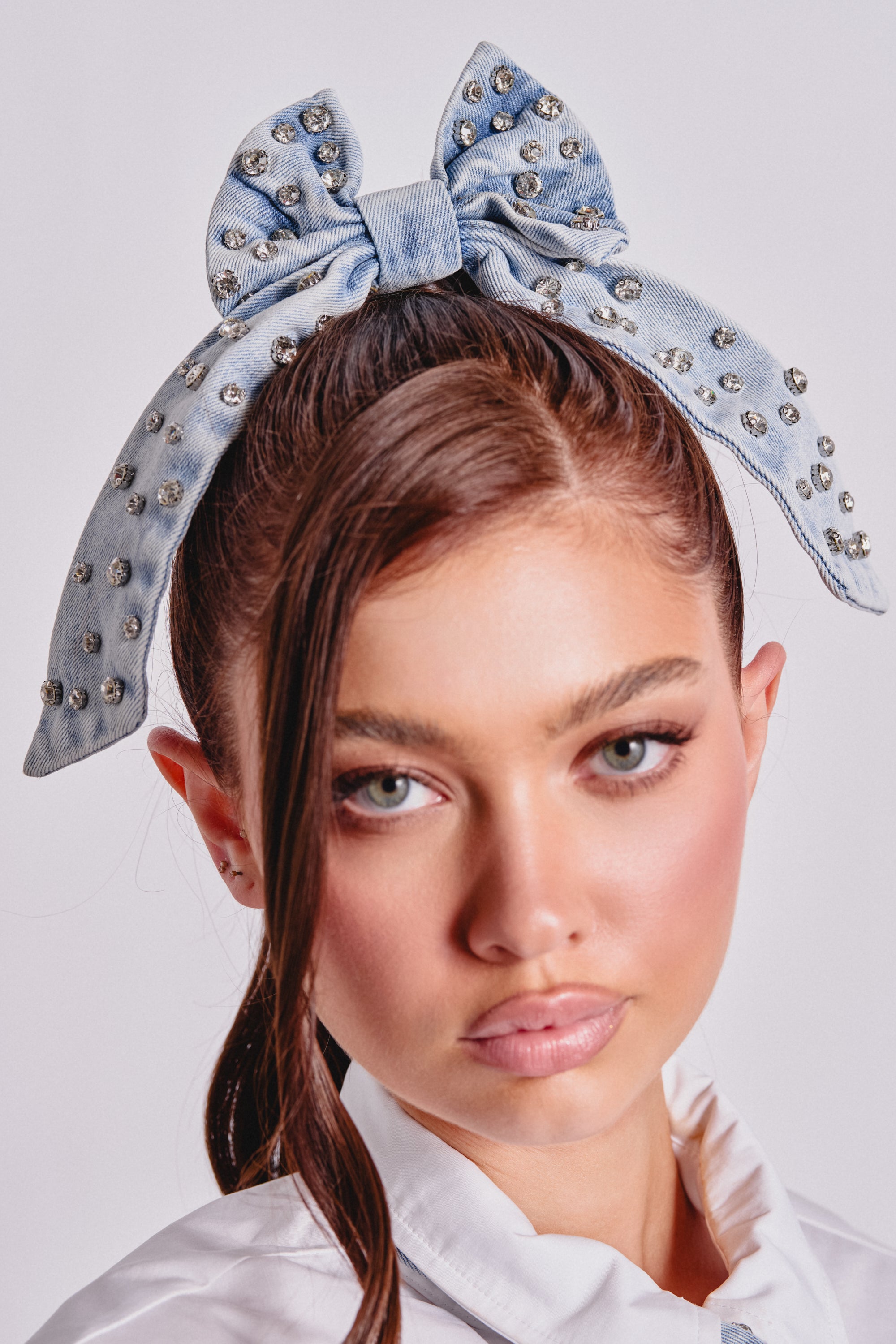 A young woman with long brown hair wears the DENIM AND DIAMONDS HAIR BOW—an oversized, light blue denim accessory adorned with silver studs—atop her head. She pairs it with flawless makeup and a white collared shirt. 1