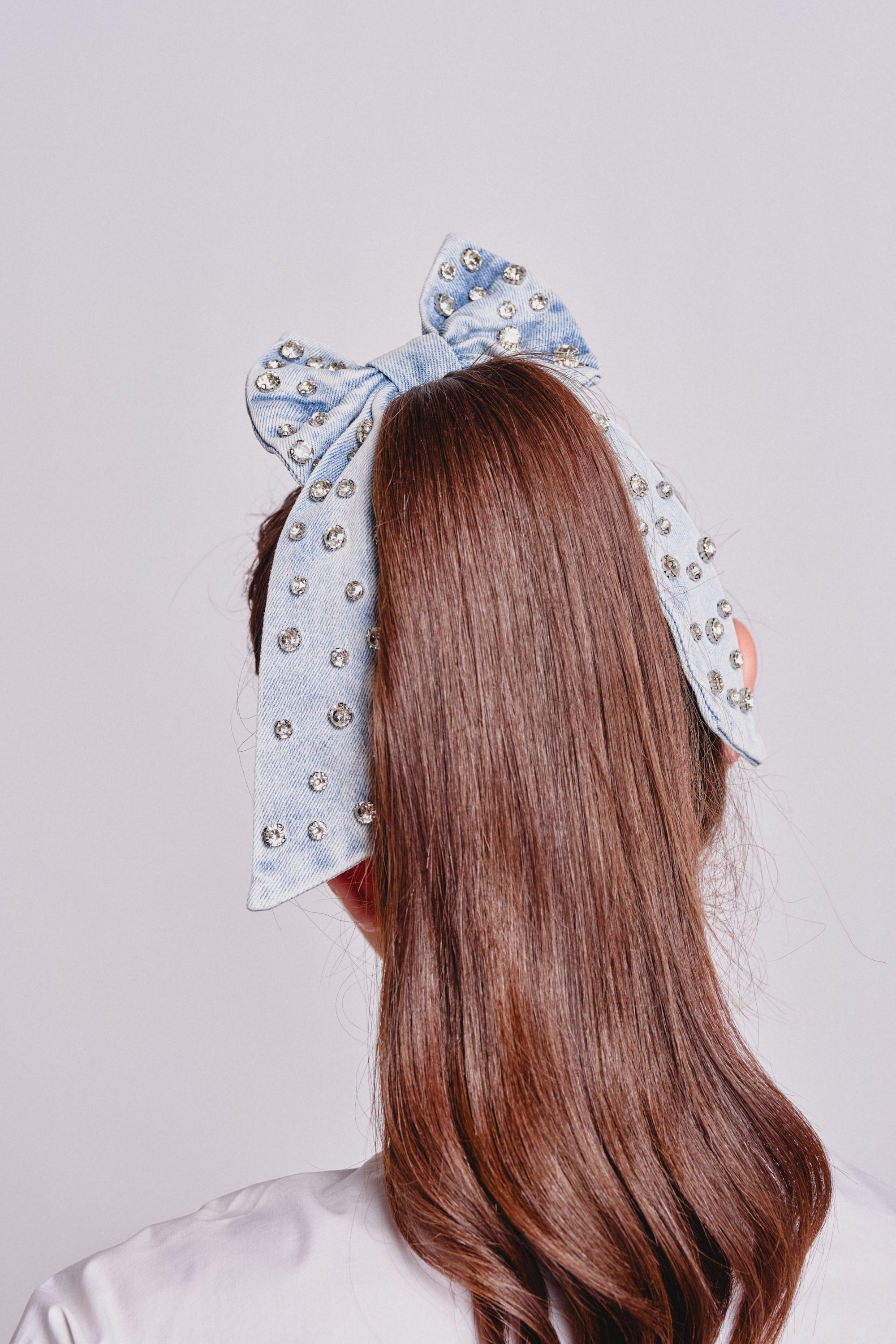 A person with long brown hair wears the DENIM AND DIAMONDS HAIR BOW, an oversized light blue denim bow adorned with sparkling rhinestones, styled at the top of their head and paired with a crisp white shirt. 4