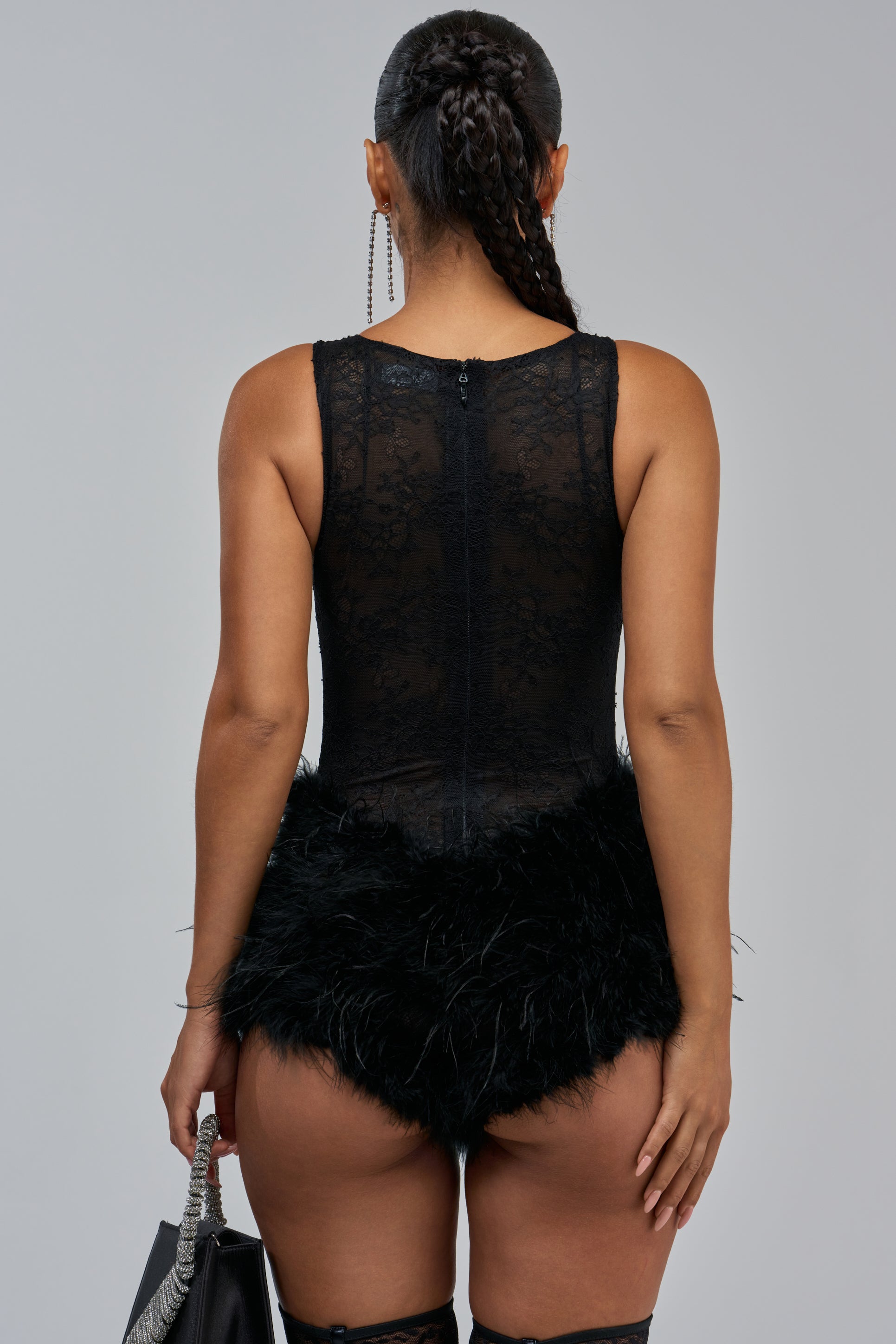 A woman stands facing away, wearing the NECESSARY DRAMA FEATHER ROMPER—a sexy one-piece black lace outfit with a feathered skirt—paired with thigh-high boots and a black handbag. Her hair is braided in a long ponytail.