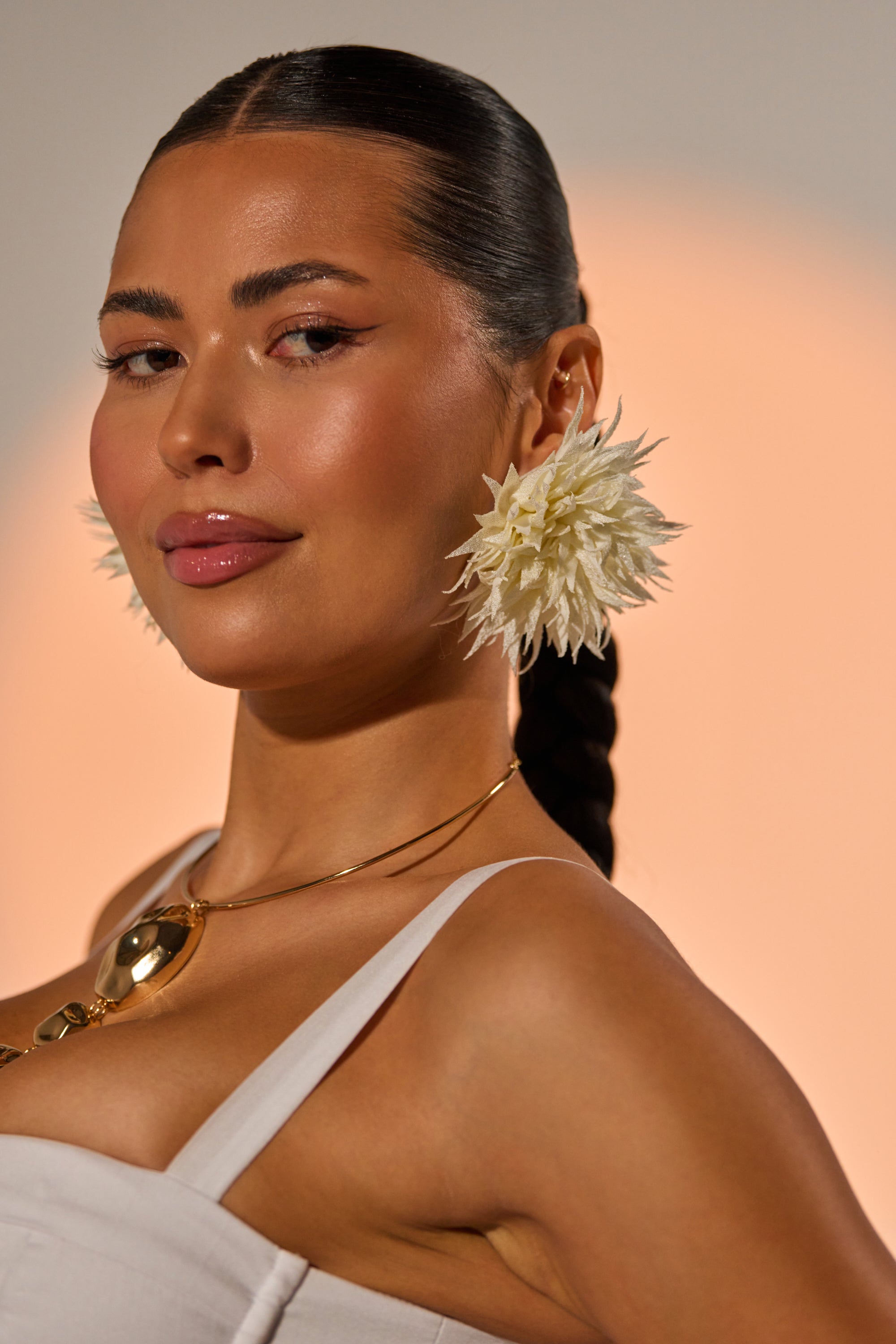A woman with sleek, dark hair in a long braid wears the LALA LAND EARRING, a gold necklace, and a white top. Her glowing skin and subtle smile stand out against a soft peach background. 0
