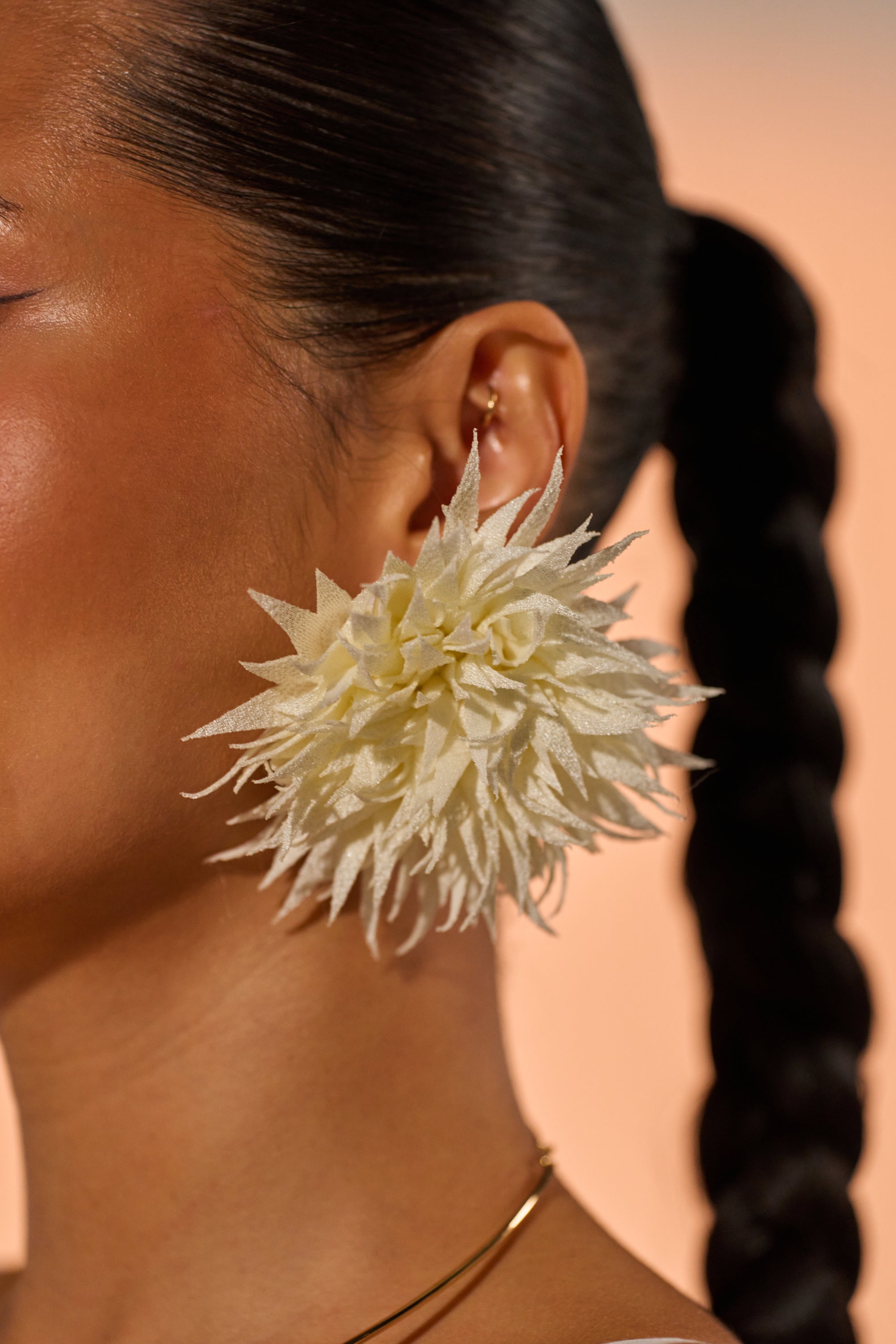 A close-up of a woman with a long braided ponytail wears the LALA LAND EARRING—large, floral-inspired statement jewelry—and a gold necklace, soft lighting highlighting her smooth skin. 1