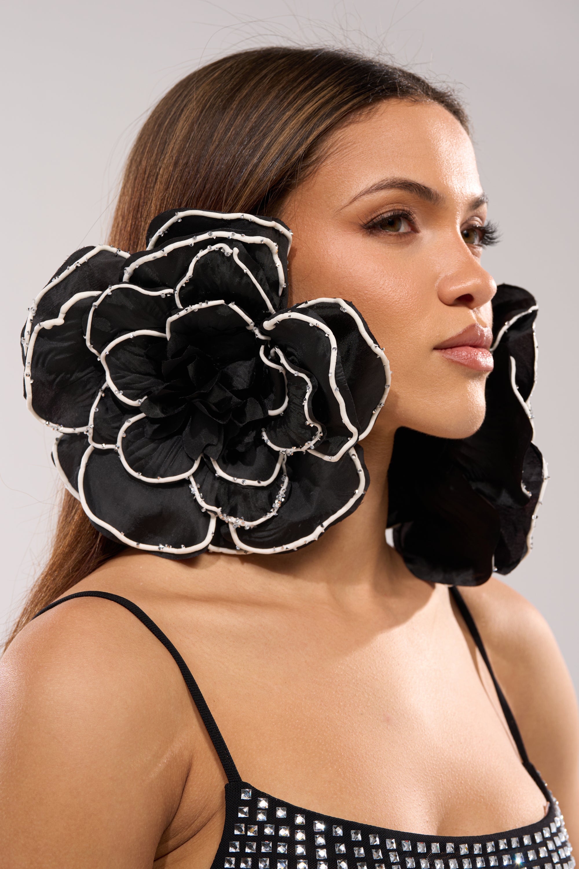 A woman with long brown hair wears the DRAMA EARRING—oversized floral earrings with blooming petals and white edges. She pairs them with a black top featuring silver studs, posing against a plain, light gray background.
