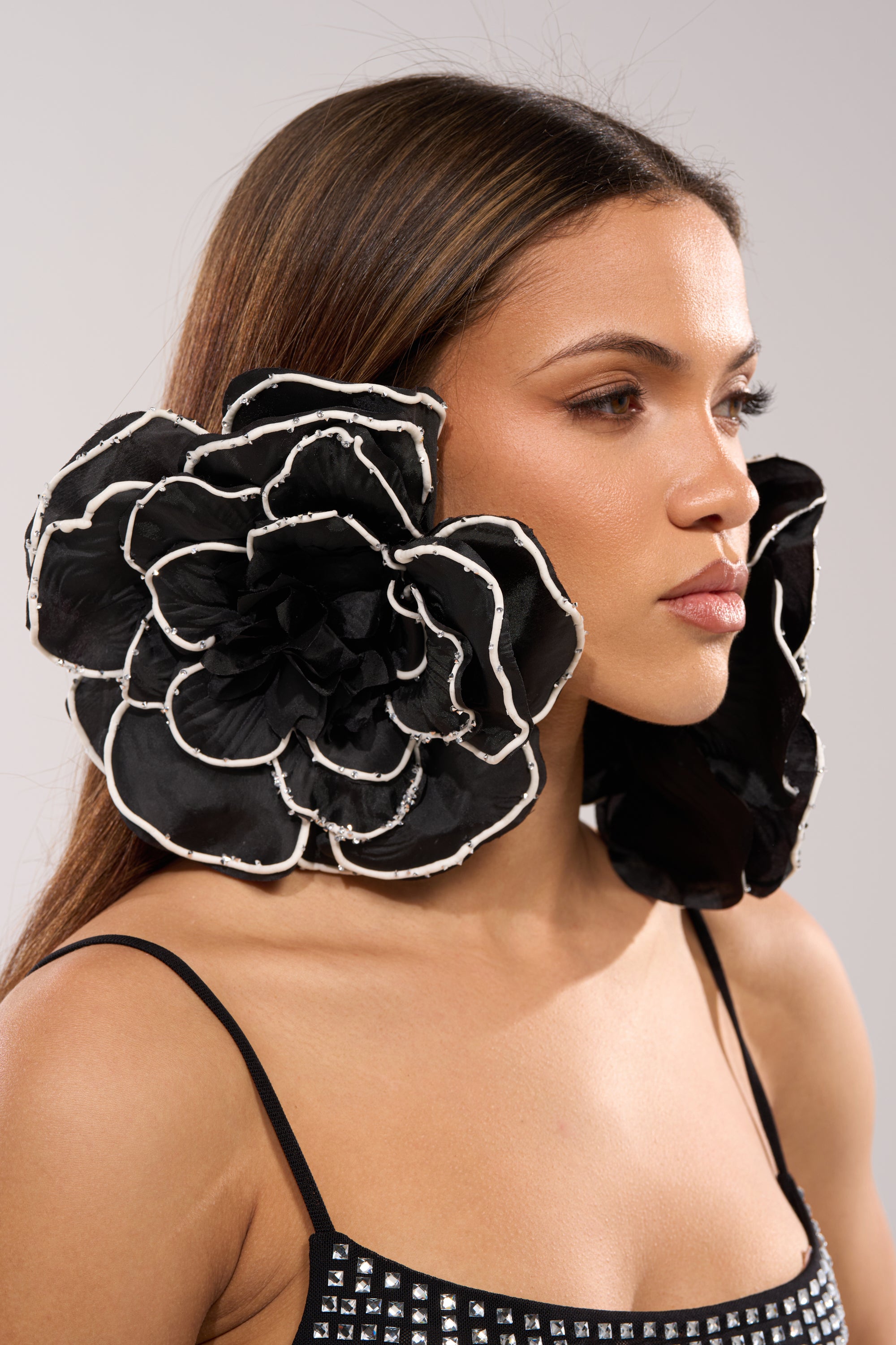 A woman with long brown hair wears DRAMA EARRING—oversized floral earrings with layered petals and white edges—paired with a black spaghetti strap top with silver studs, posing against a neutral background.
