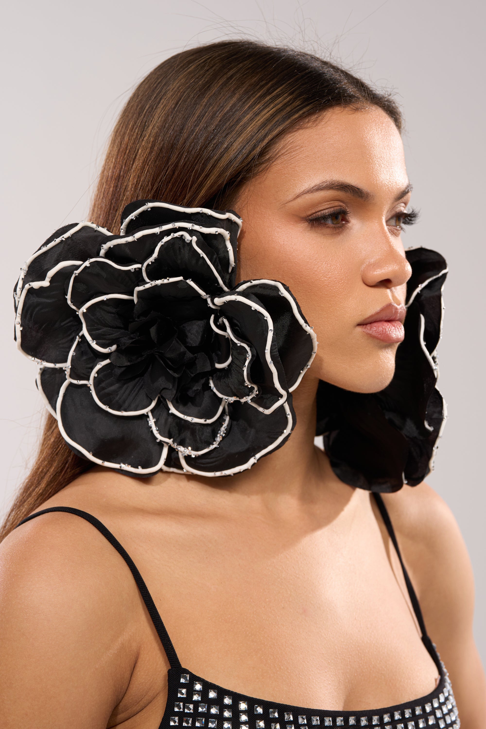 A woman with long brown hair wears DRAMA EARRING—oversized floral earrings with layered petals and white edges—paired with a black spaghetti strap top with silver studs, posing against a neutral background. 1