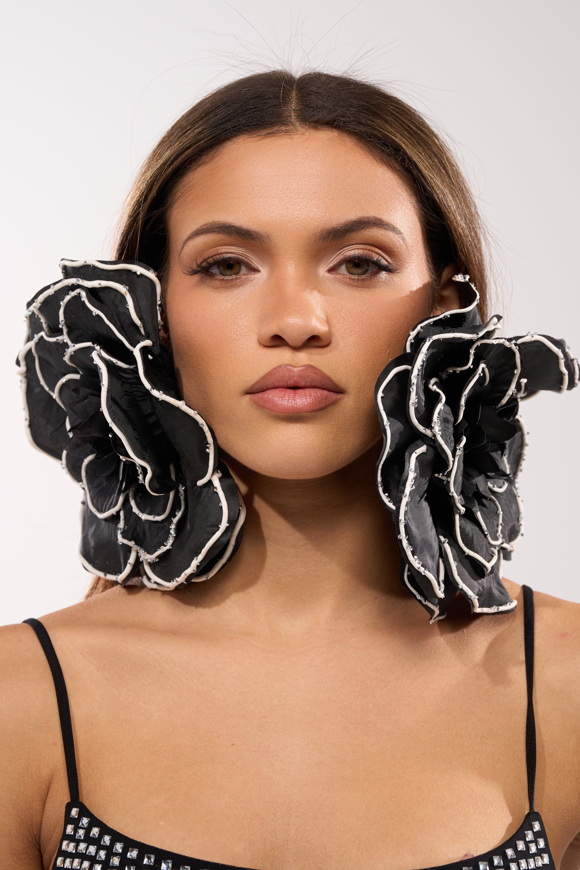 A woman with straight brown hair wears DRAMA EARRING—oversized black and white floral statement earrings—framing her face. She has a neutral expression and is dressed in a black spaghetti strap top against a plain, light background. 2
