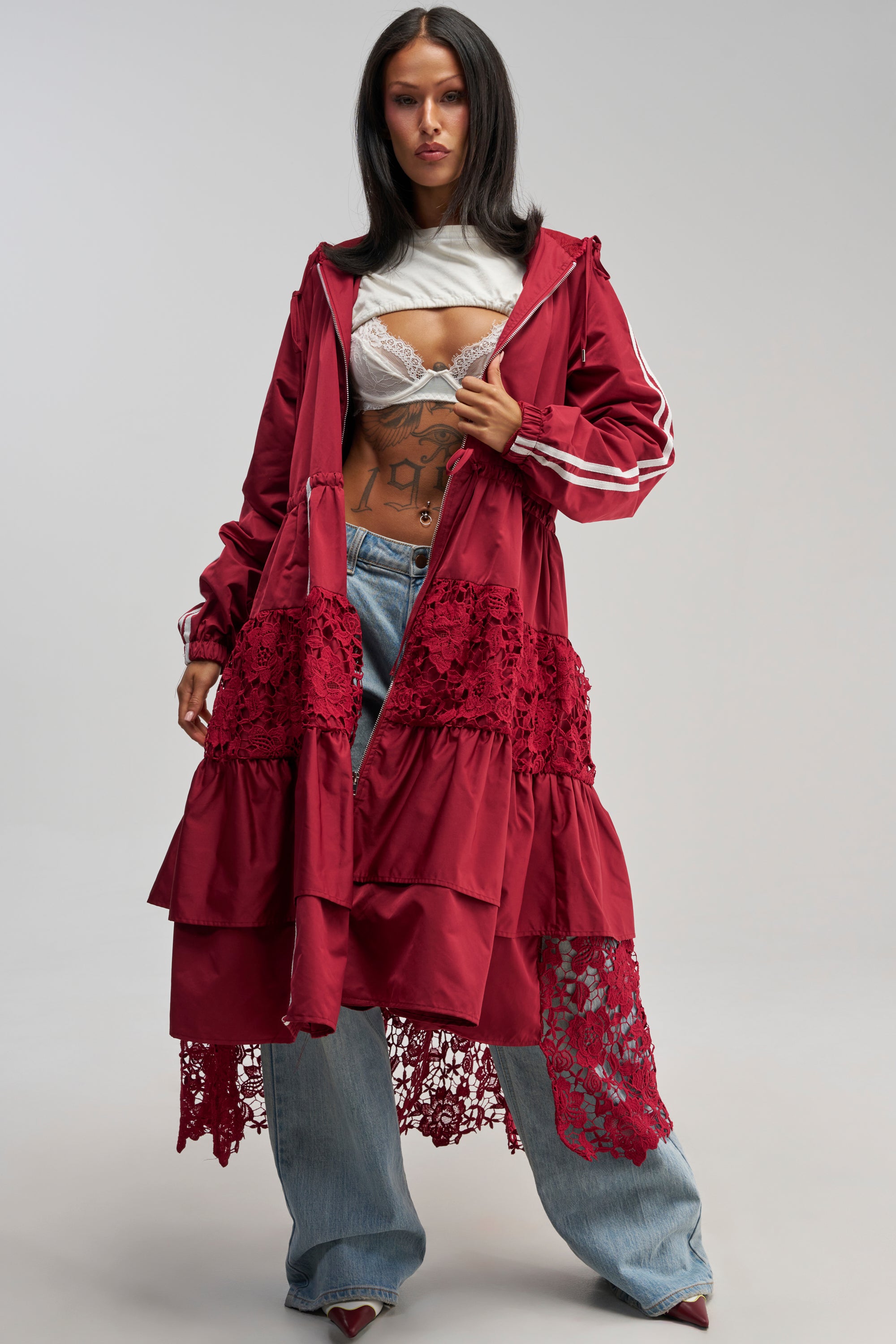 A woman wears the READY FOR IT TRENCH, a long burgundy trench coat with intricate lace detailing, layered over a white lace bralette, paired with loose blue jeans and pointed red shoes, while standing against a plain gray background.