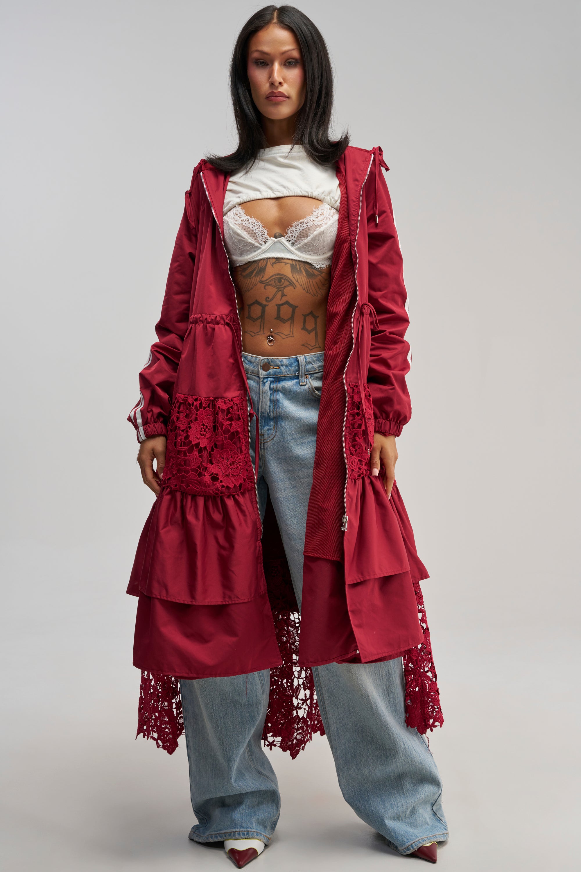 A woman wears the READY FOR IT TRENCH, a long burgundy coat featuring lace detailing, styled over a white bra and top with loose blue jeans. She has long dark hair, tattoos visible on her stomach, and stands against a plain light background.
