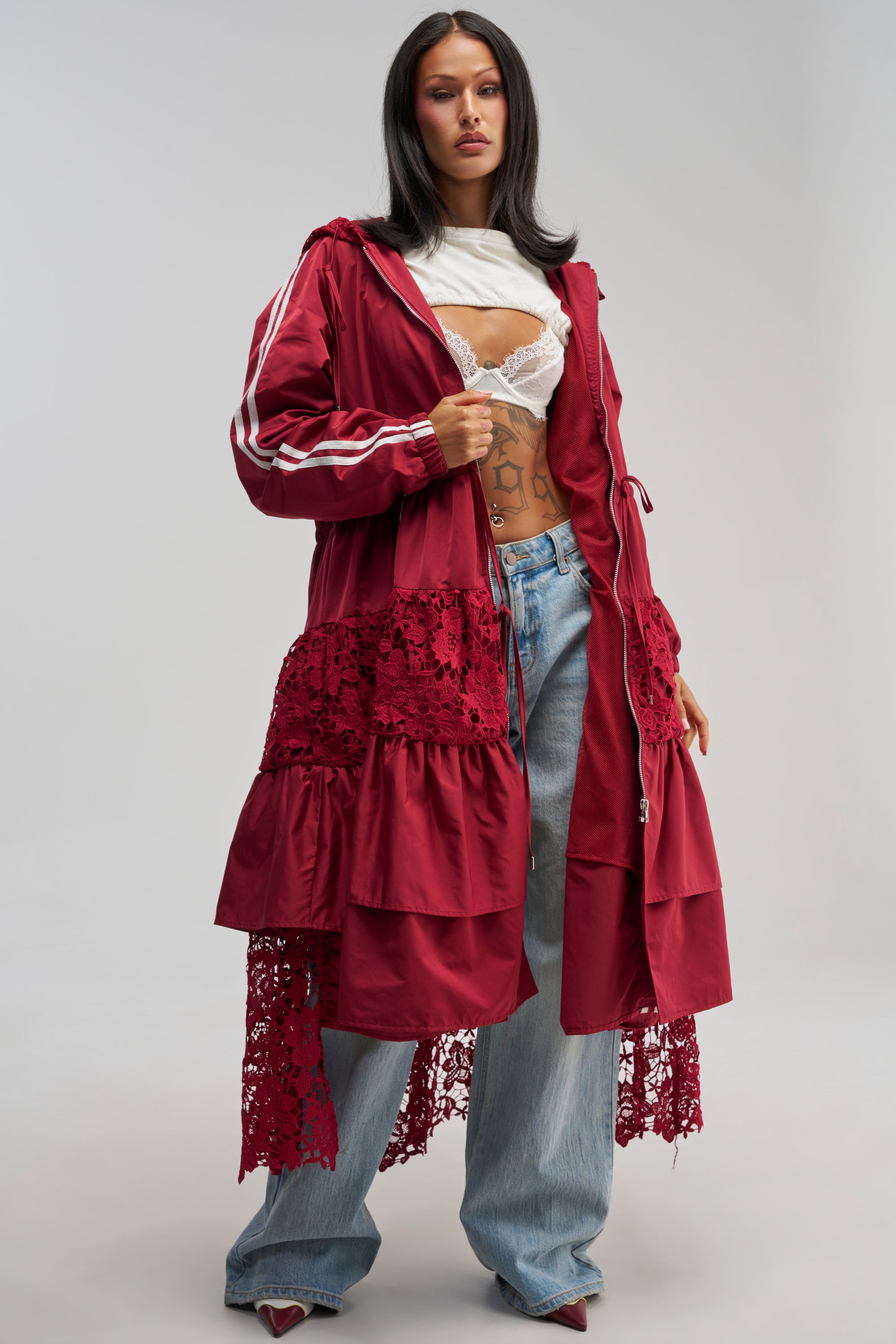 Wearing the READY FOR IT TRENCH, a long burgundy coat with lace detailing, styled with a white lace bralette, loose blue jeans, red pointed shoes, and a white scarf around her neck, the woman stands confidently against a plain gray background.