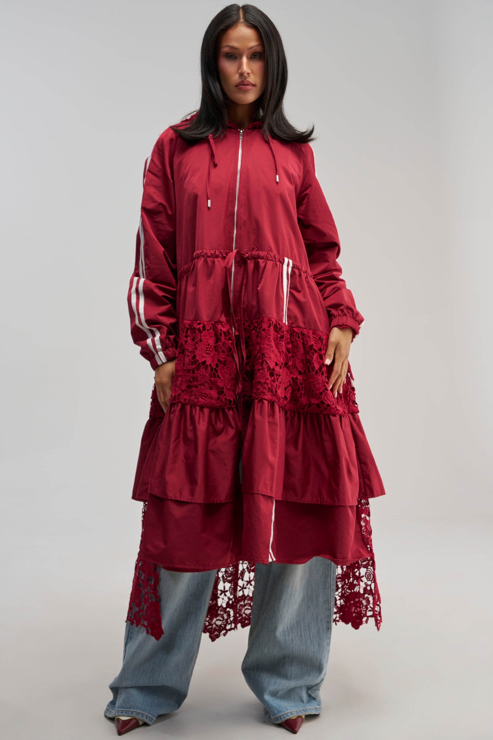 Wearing the READY FOR IT TRENCH, a long maroon streetwear jacket with lace and ruffle details, over light blue jeans and pointed shoes, a person stands against a plain light background, looking directly at the camera.