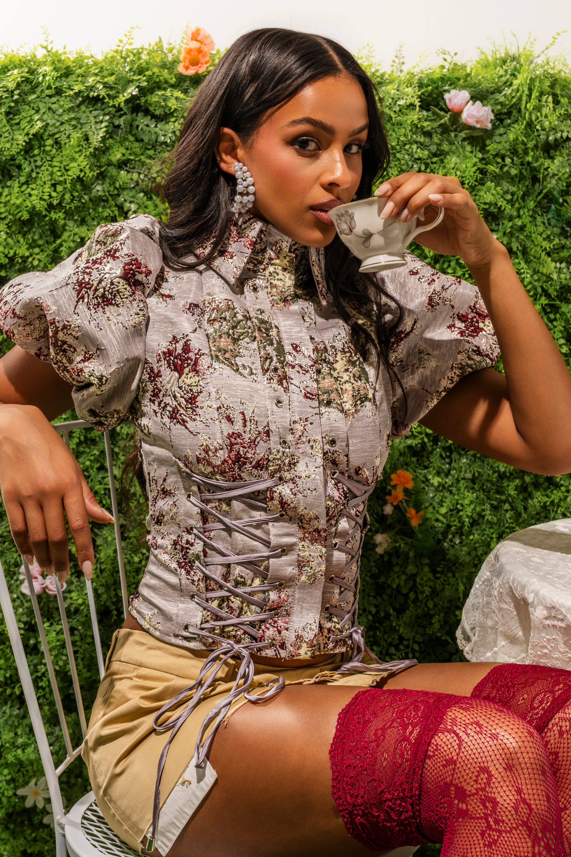 A woman with long dark hair sips tea from a delicate cup, wearing the DARLING FLORAL BROCADE PUFF SLEEVE BLOUSE and a beige skirt, seated by a garden table surrounded by lush foliage and blooming flowers. 0