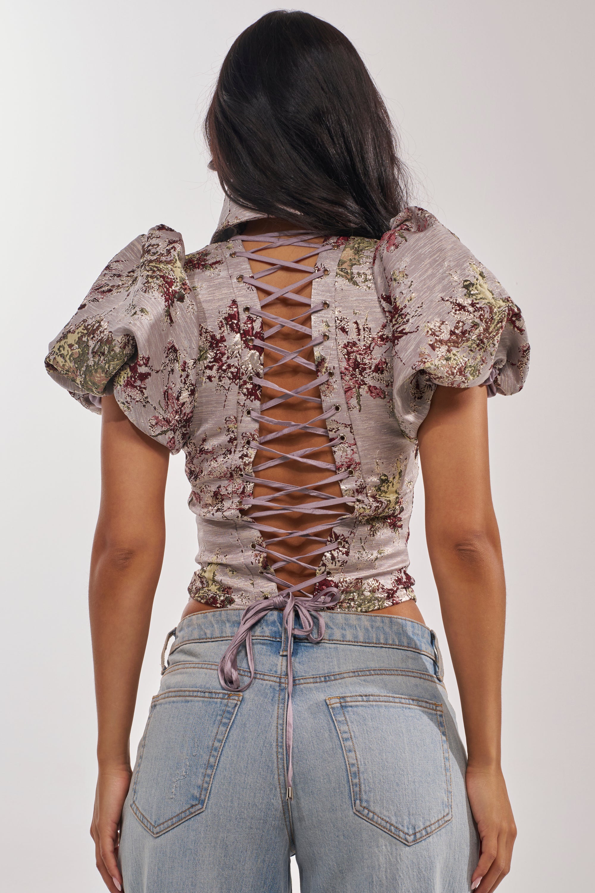 A woman with long dark hair is seen from the back, wearing the DARLING FLORAL BROCADE PUFF SLEEVE BLOUSE with a corset-inspired lace-up back and vintage fit, paired with light blue jeans. 3