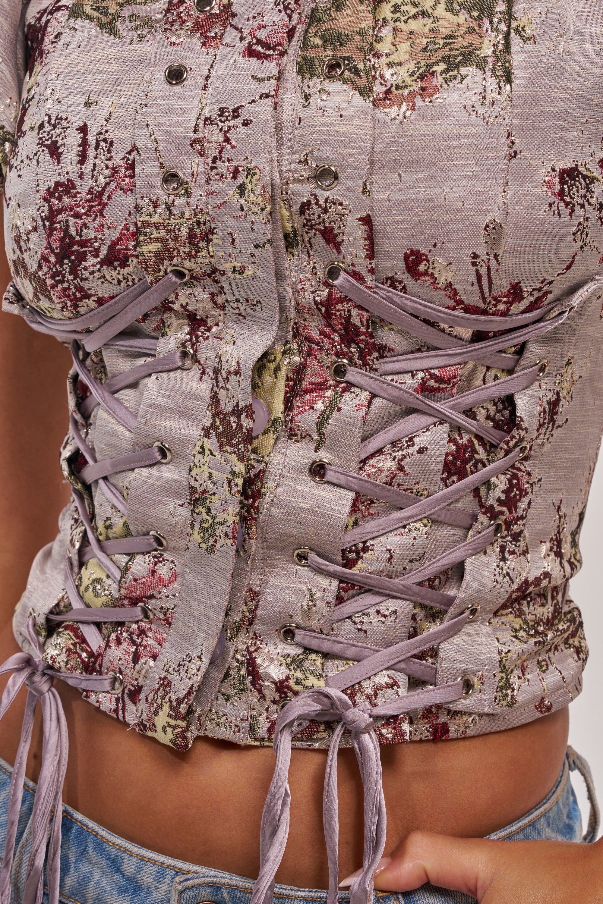 Close-up of a person wearing the DARLING FLORAL BROCADE PUFF SLEEVE BLOUSE, featuring corset-inspired crisscrossed lace-up front with metal eyelets, textured floral pattern, and paired with light blue denim jeans. 6