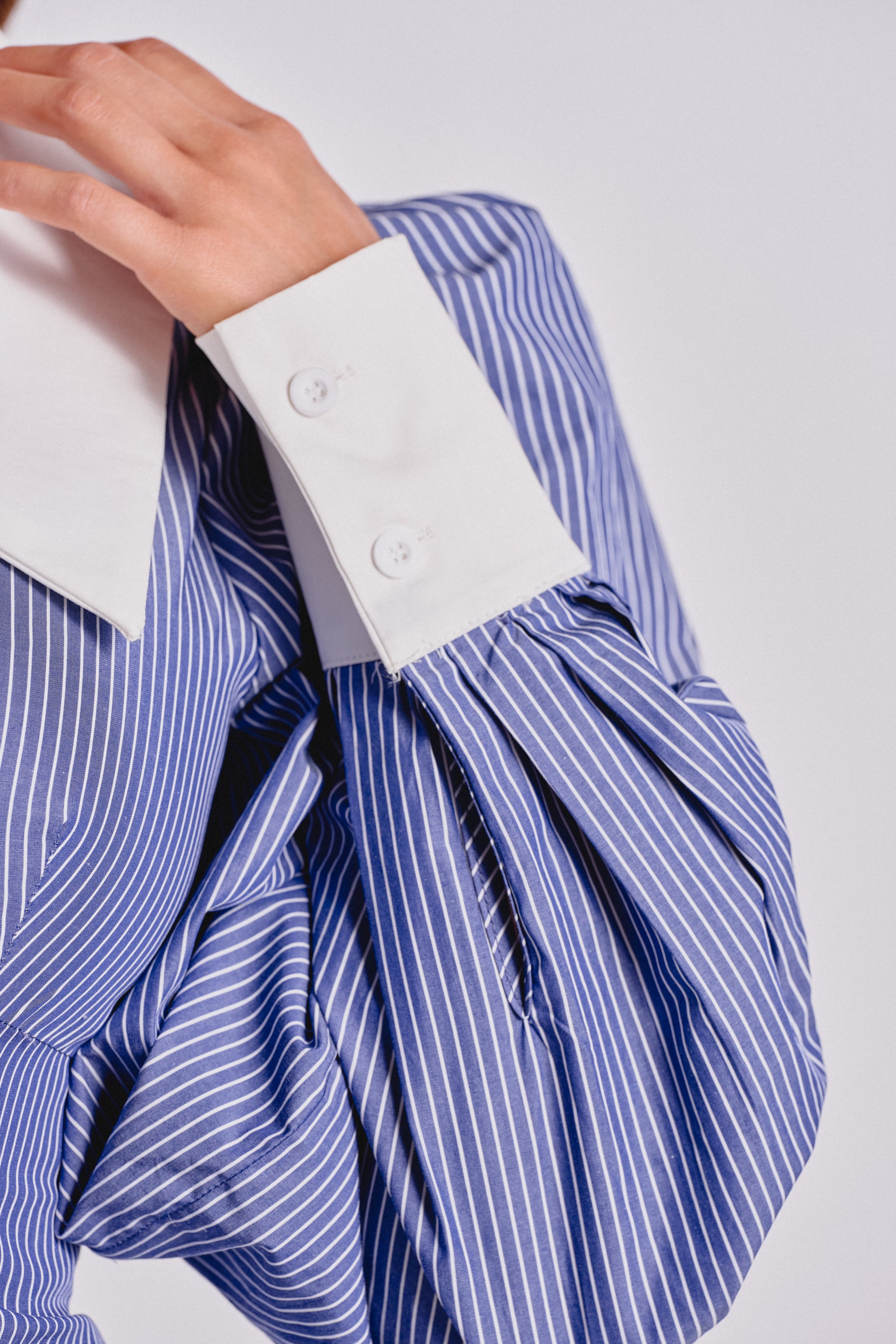 A close-up of a person in the CHELSEA LONG SLEEVE STRIPE MINI DRESS, featuring blue pinstripes, voluminous sleeves, wide white collar, and large buttoned cuffs, adjusting the collar for a crisp, polished look. 5