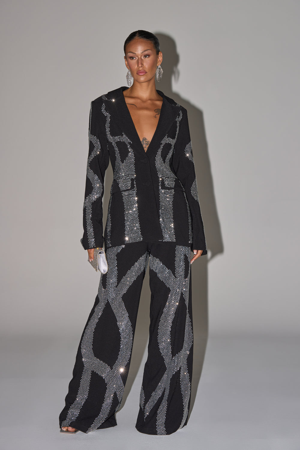 A woman poses in GEMINI RHINESTONE PANT—bold black wide-leg trousers adorned with large, wavy silver sequins. She pairs them with a deep-neckline blazer, statement earrings, and a small silver clutch.