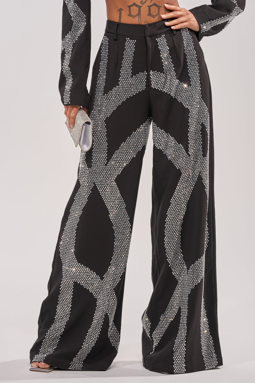 Wearing the GEMINI RHINESTONE PANT with bold silver geometric rhinestones, a person holds a small silver clutch, hand on hip, revealing a lower abdomen tattoo.
