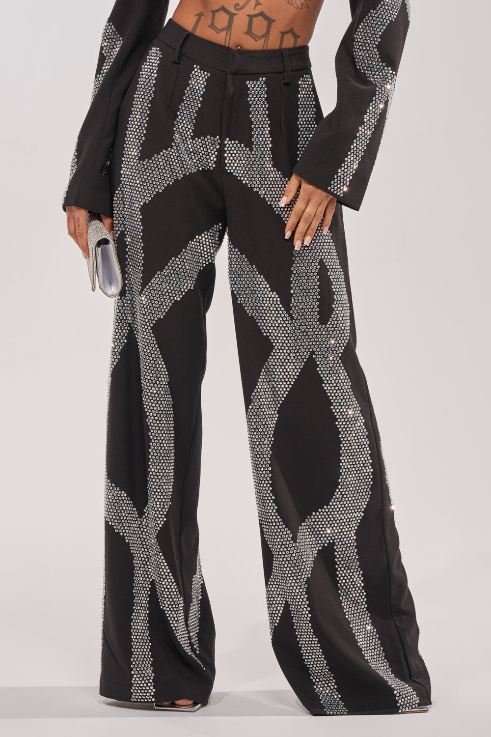 Wearing the GEMINI RHINESTONE PANT—high-rise black trousers with silver sequin swirl patterns—and holding a small silver clutch, this person shows their midriff and part of a matching jacket.
