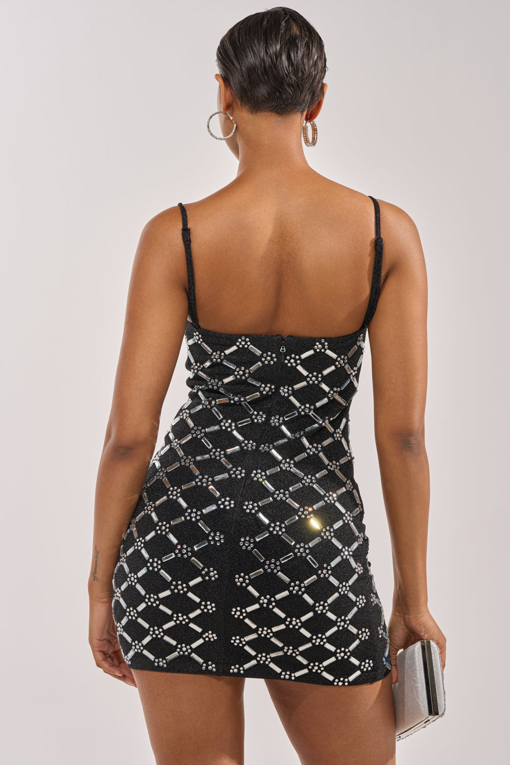 A person with short hair is seen from the back wearing the SPARKLE CITY RHINESTONE MINI DRESS—a fitted black party dress with a geometric white pattern. They hold a small clutch and wear large hoop earrings. 7