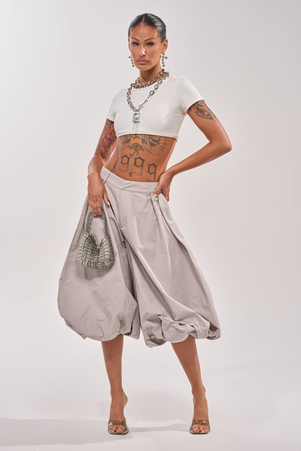 Model stands confidently in a white crop top and the GLOW GAUCHO PANT—grey, high-waisted with an elastic waistband—paired with clear heels, a silver handbag, bold statement jewelry, and striking arm and torso tattoos. 0