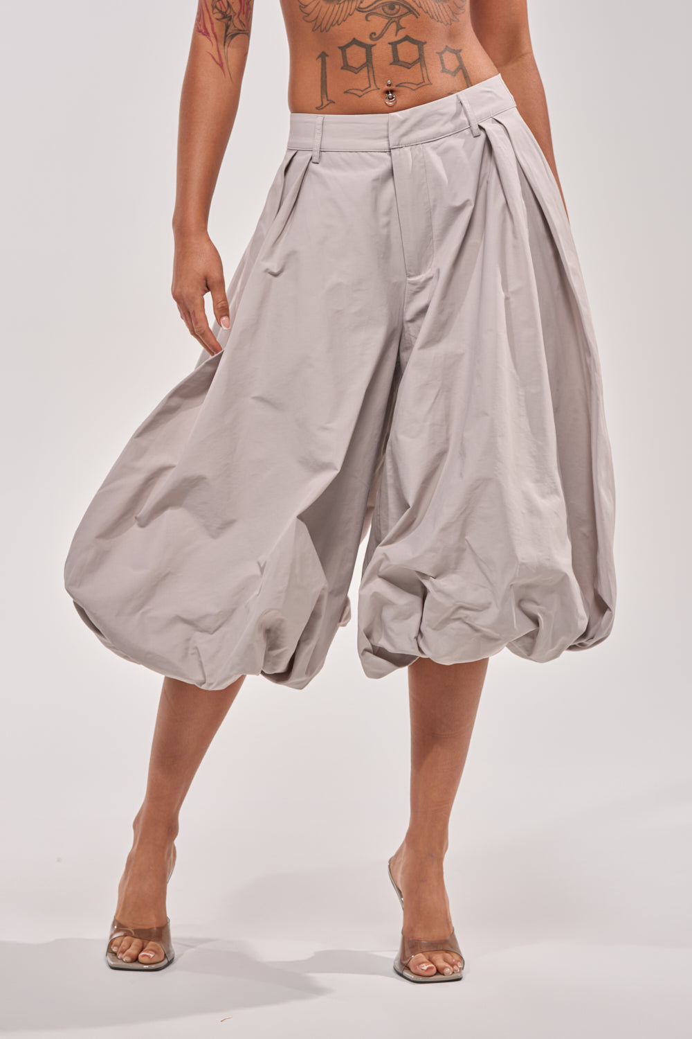 A person wears the GLOW GAUCHO PANT in light gray with loose, pleated detailing and balloon-style shape, paired with clear high heels; their tattooed torso is visible against a white backdrop—ideal for modern daywear. 1